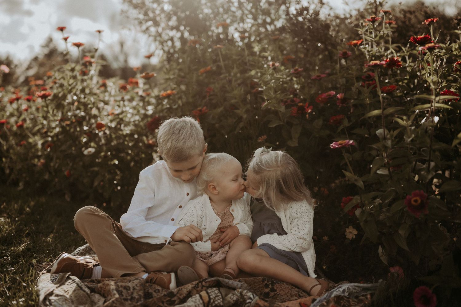 Three people share an intimate moment among flowering bushes at golden hour.