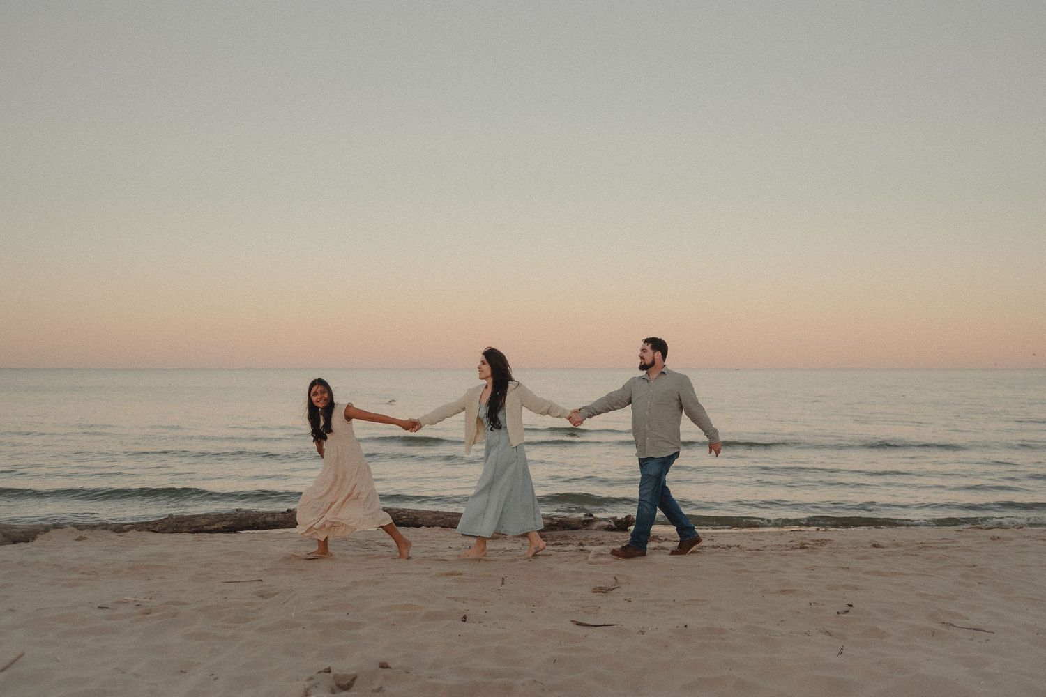 Family of three holding hands walking along beach shoreline at pink and purple dusk sunset.