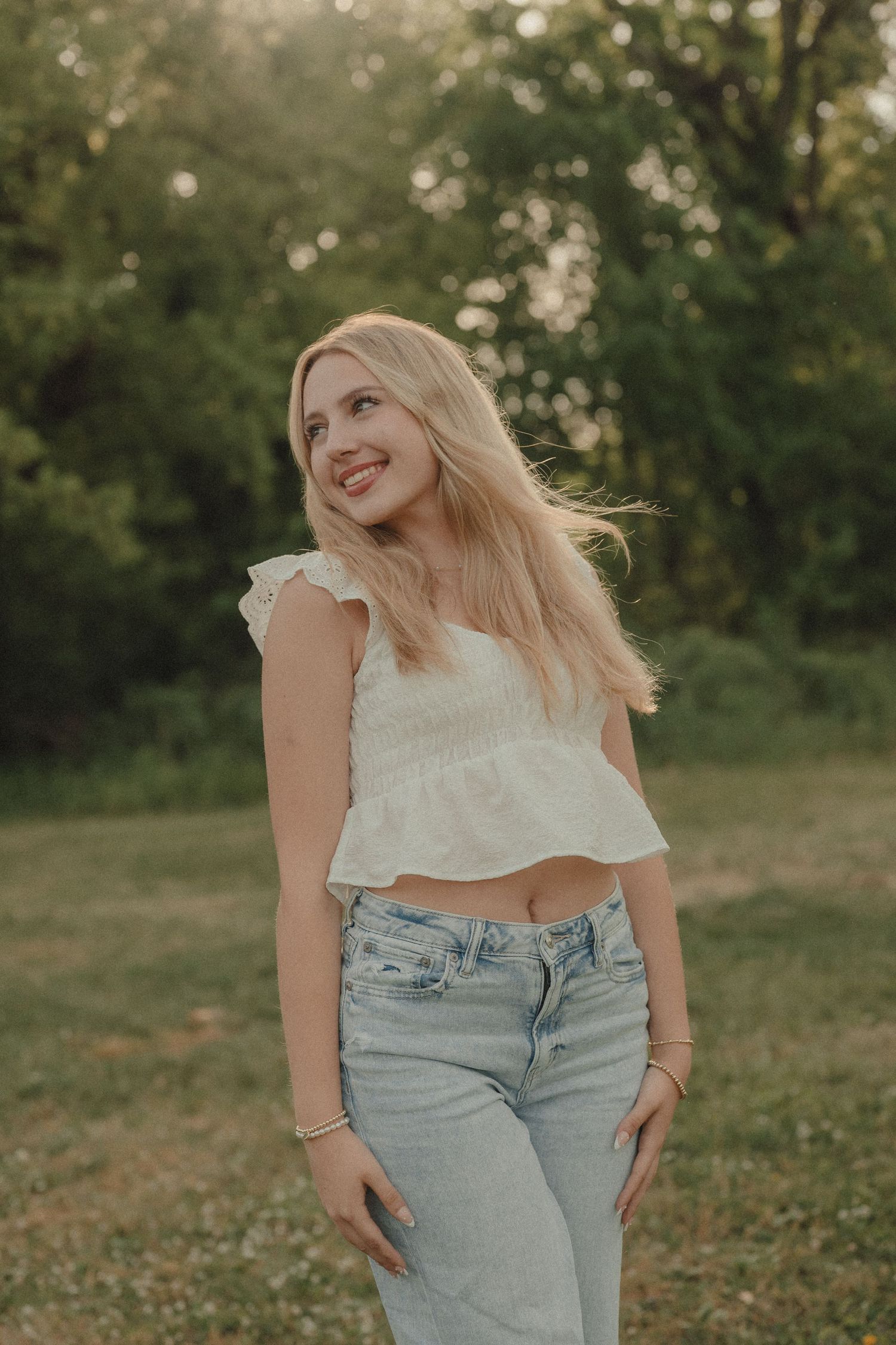 Smiling blonde woman in white crop top and jeans standing outdoors in green park at golden hour.