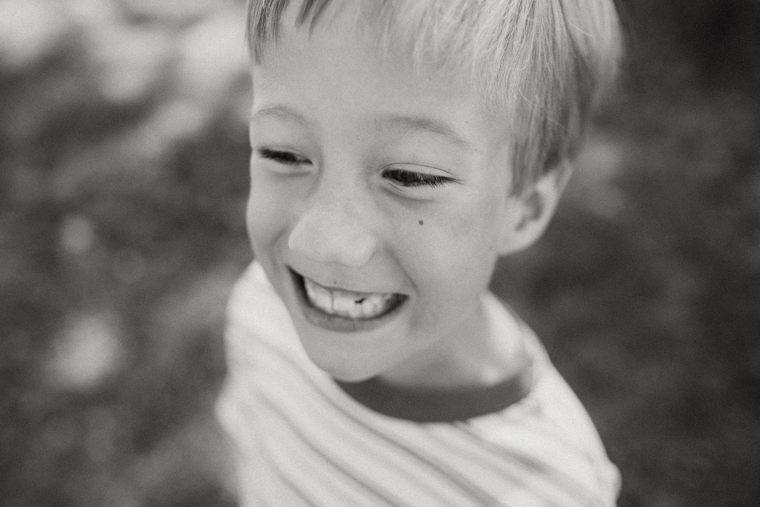 Black and white portrait of a smiling young blonde boy with a freckle on his cheek outdoors.
