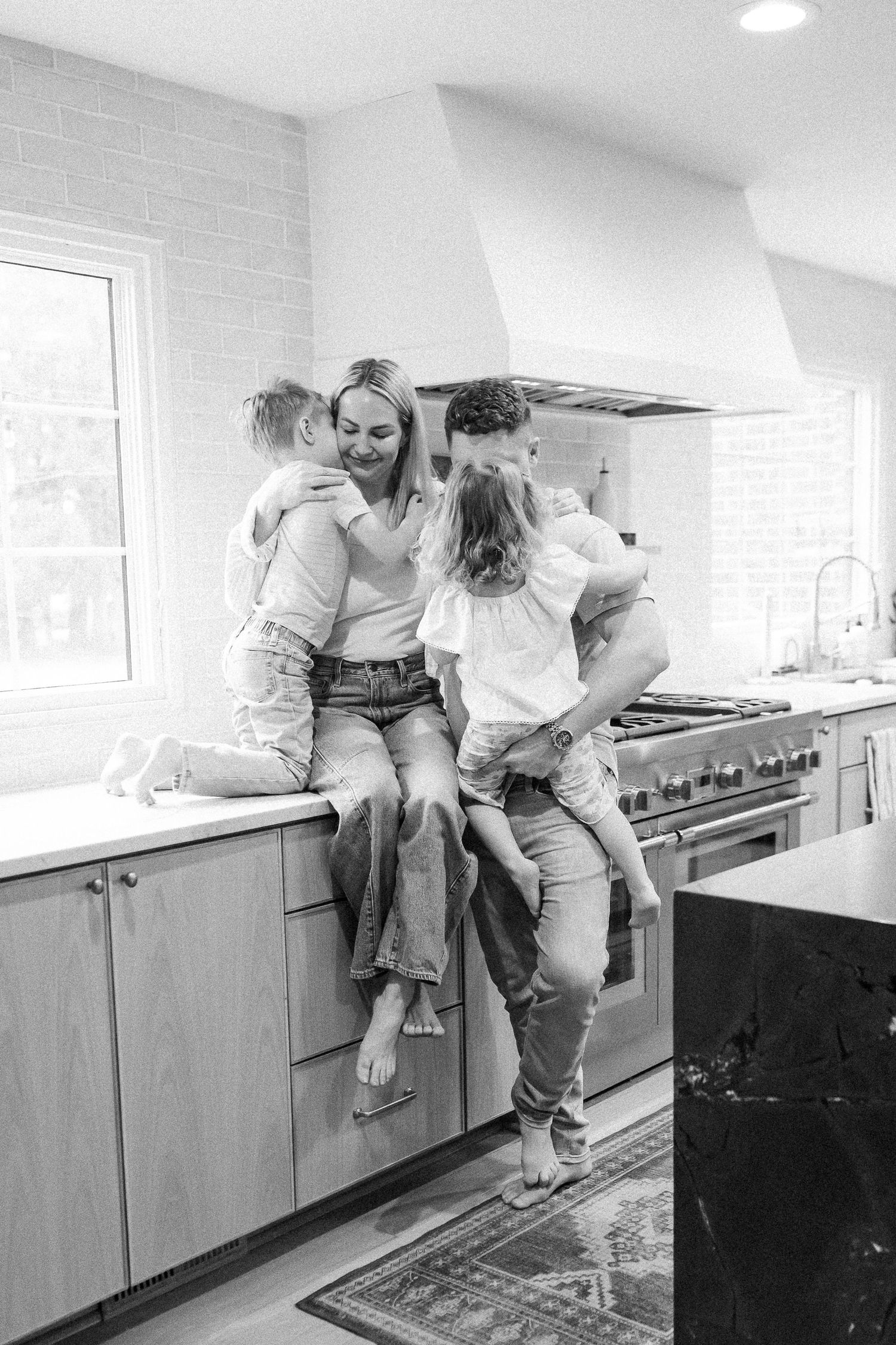 Black and white photo of a couple embracing their two young children on a kitchen counter.