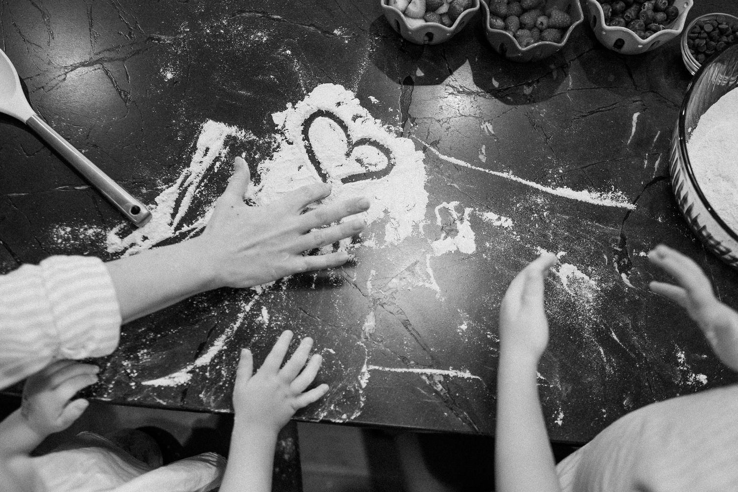 Overhead black and white photo of children's hands playing with flour on a marble countertop.