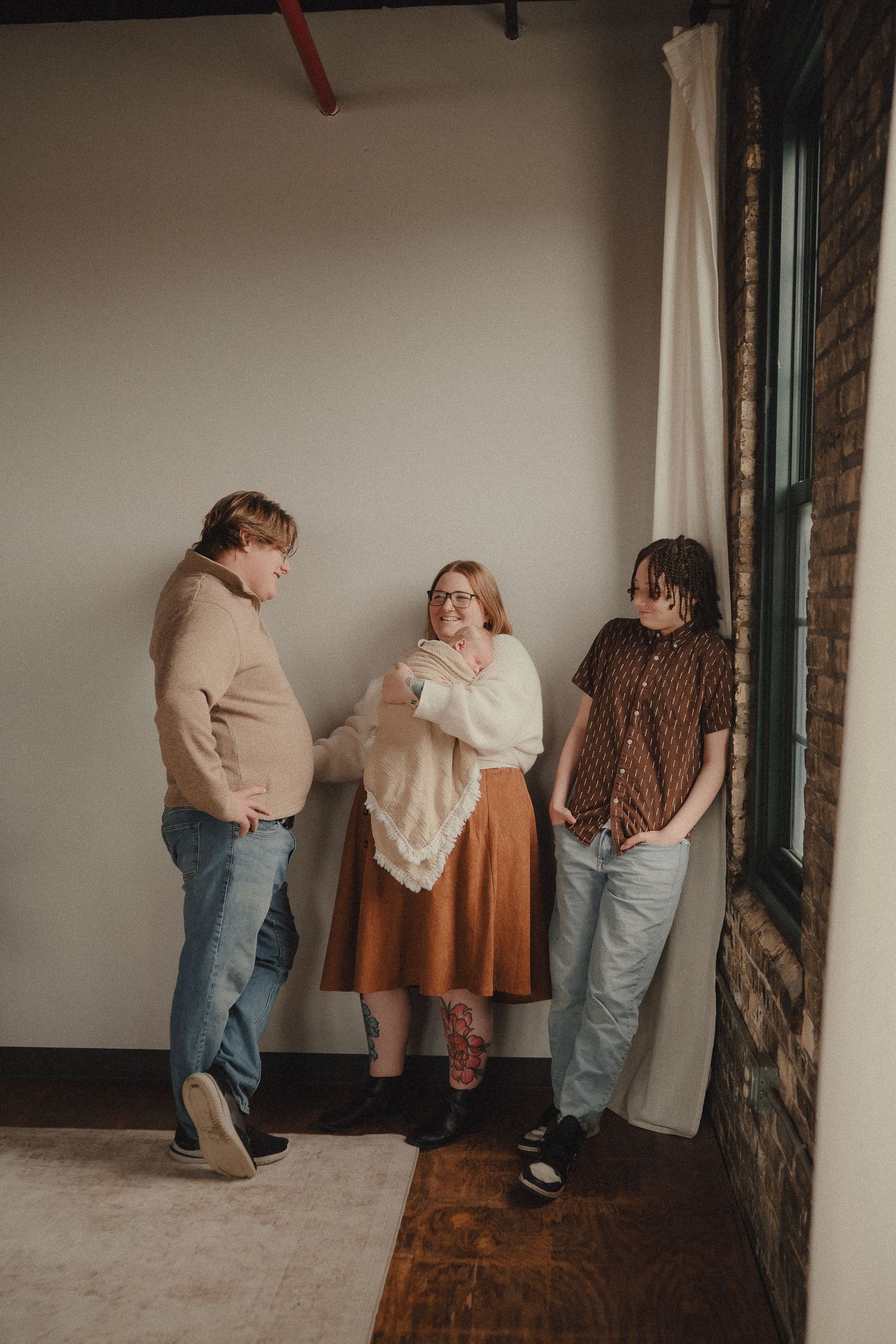 Three people standing near a window and brick wall, with a woman in a rust skirt holding a newborn baby.