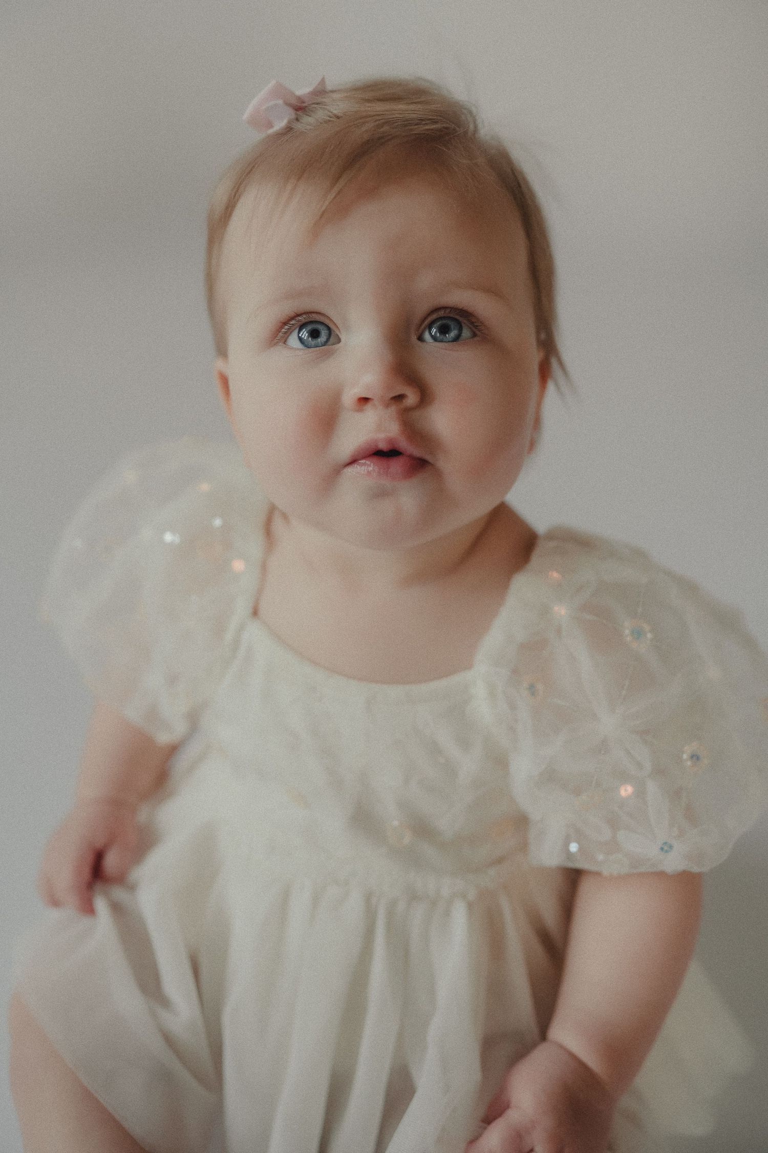 Adorable baby girl with blue eyes wearing a white sequin tulle dress with puff sleeves and pink bow hair clip.