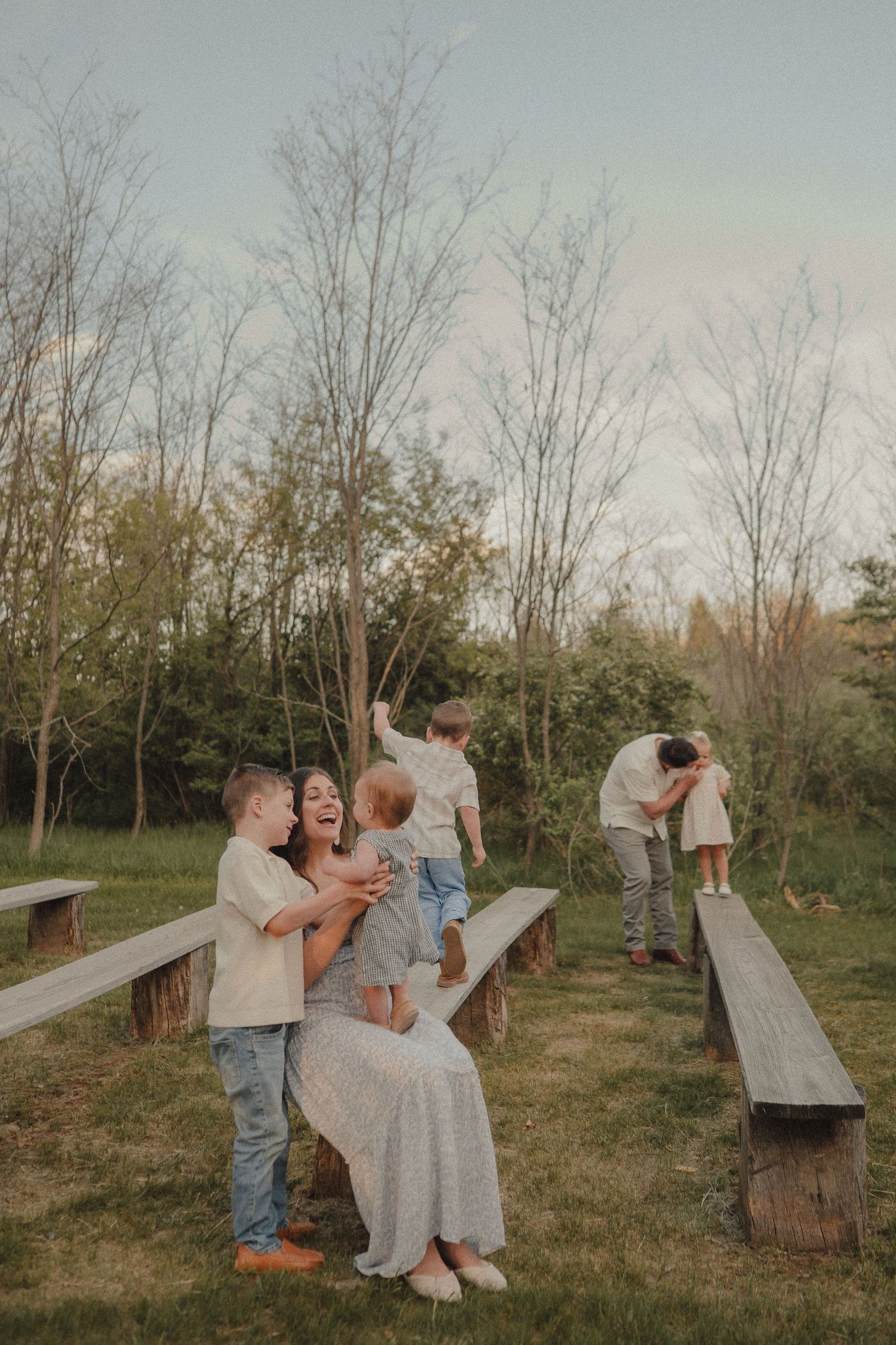 Mother laughing with children on wooden benches in a park while father plays with daughter.