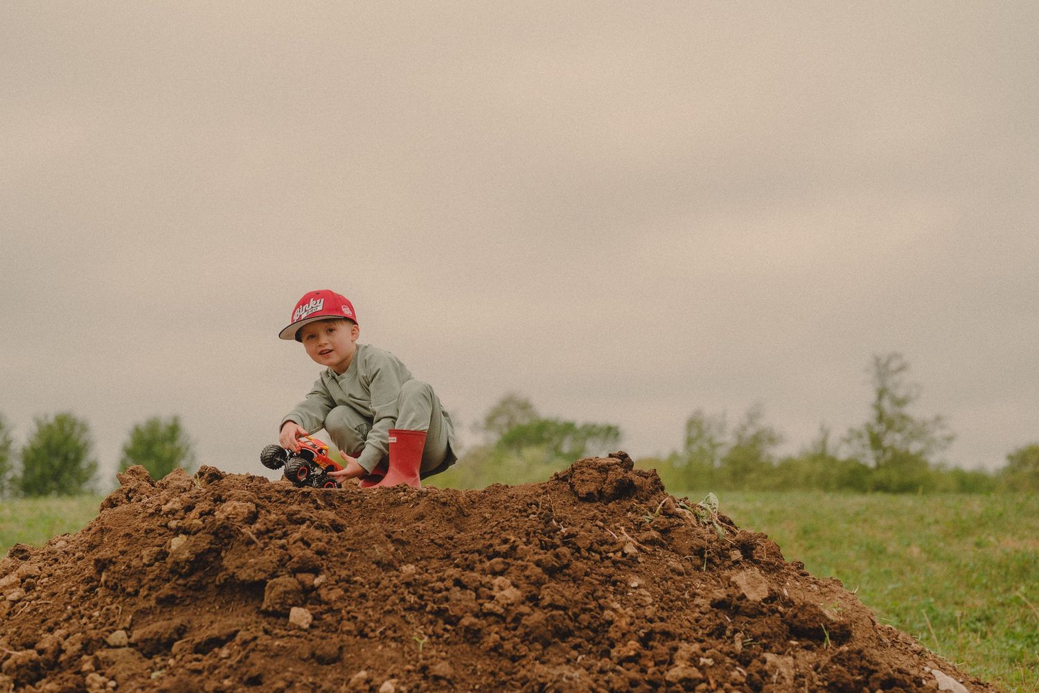 Little boy in red baseball cap crouches on pile of dirt playing with toy truck in green field.