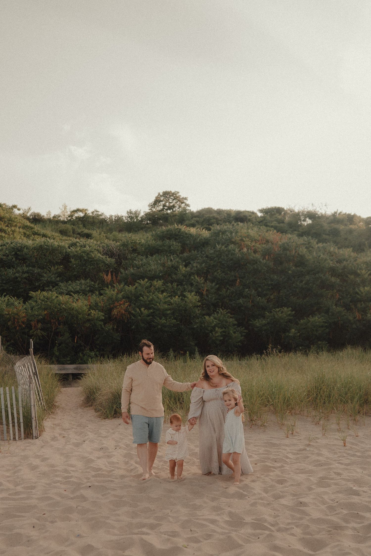 Family of four walking on sandy beach path with lush green dunes, parents holding toddler hands in neutral tones.