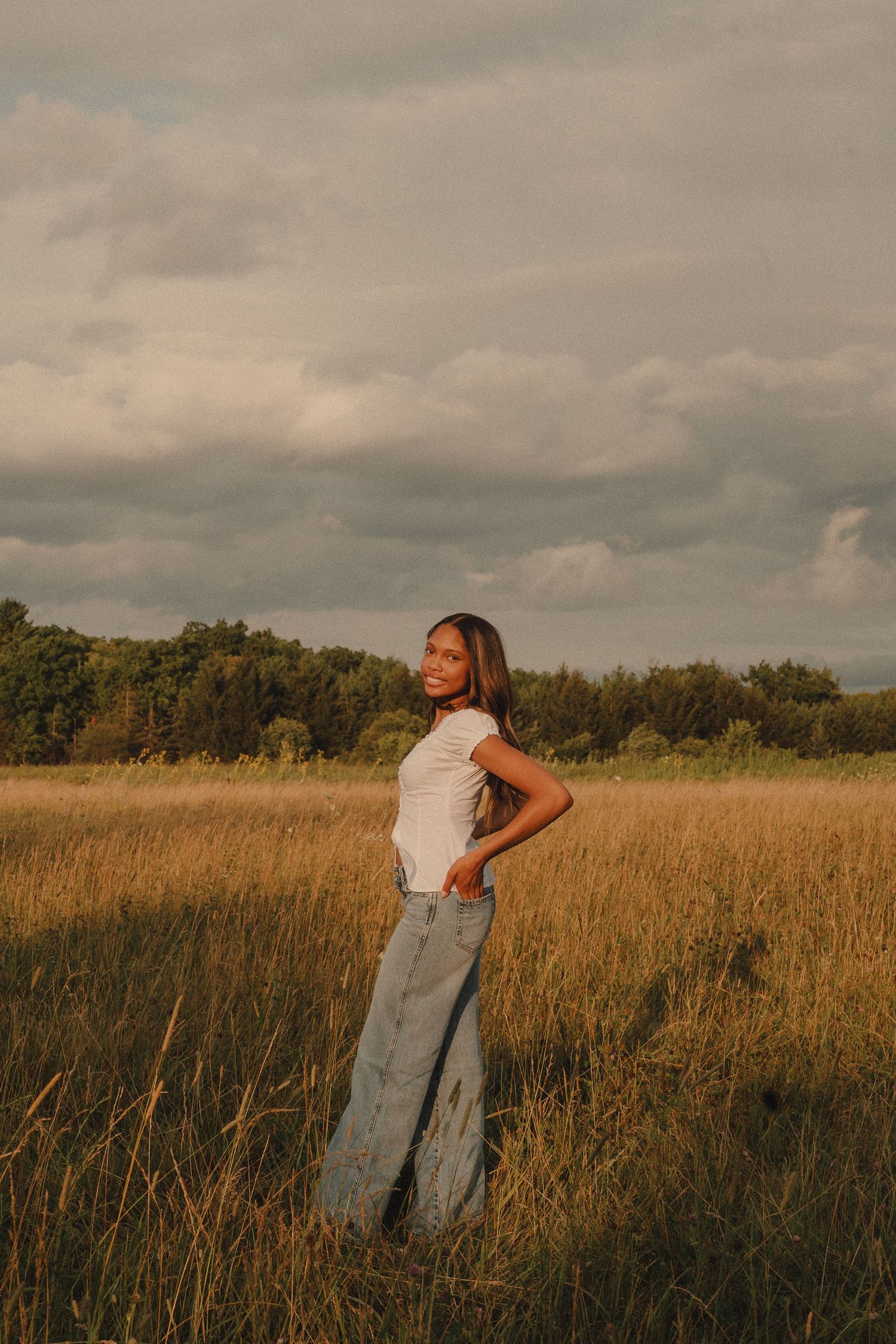 Young woman in white top and wide-leg jeans posing in a golden field under dramatic cloudy sky.