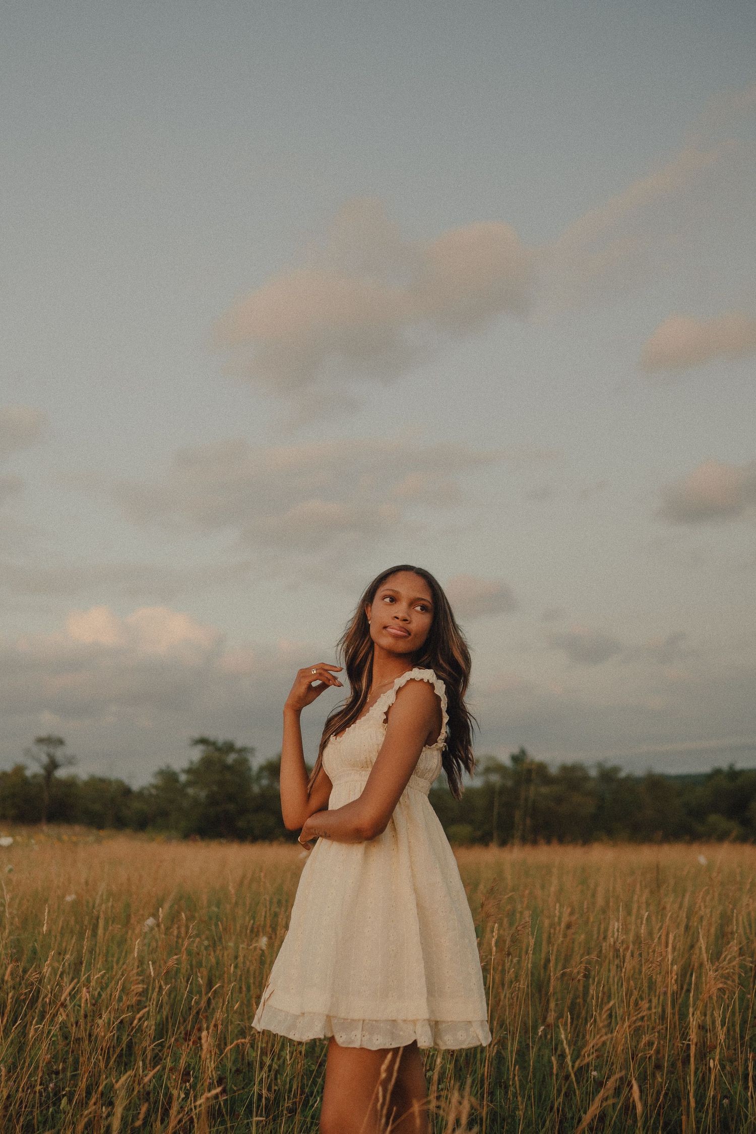 Young woman in white sundress stands in golden meadow field under a cloudy sky at dusk.