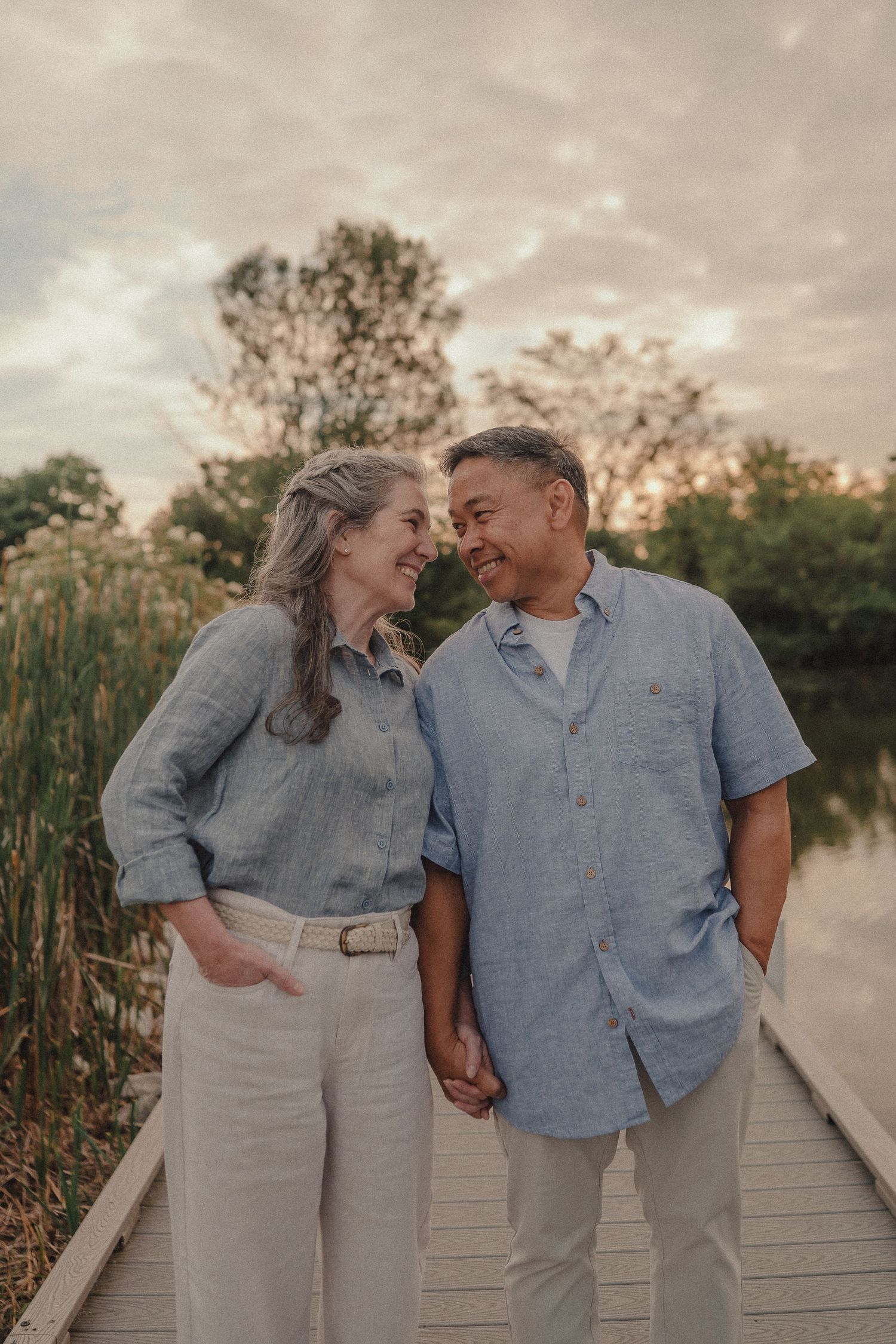 Smiling interracial couple holding hands on a wooden dock at sunset near a lake.
