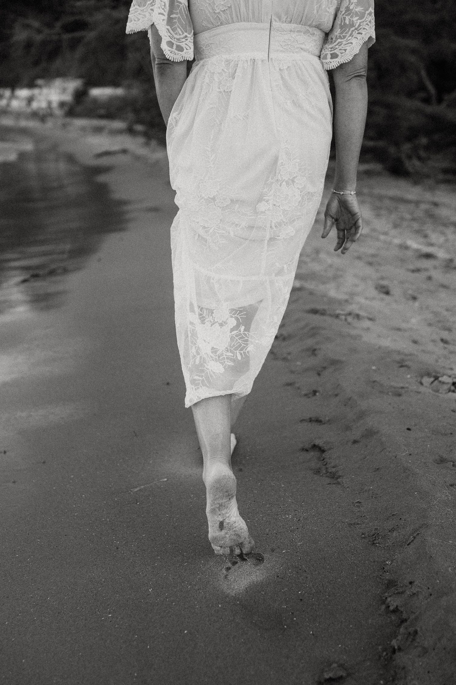 Woman in white lace dress walking barefoot on sandy beach, black and white photography.