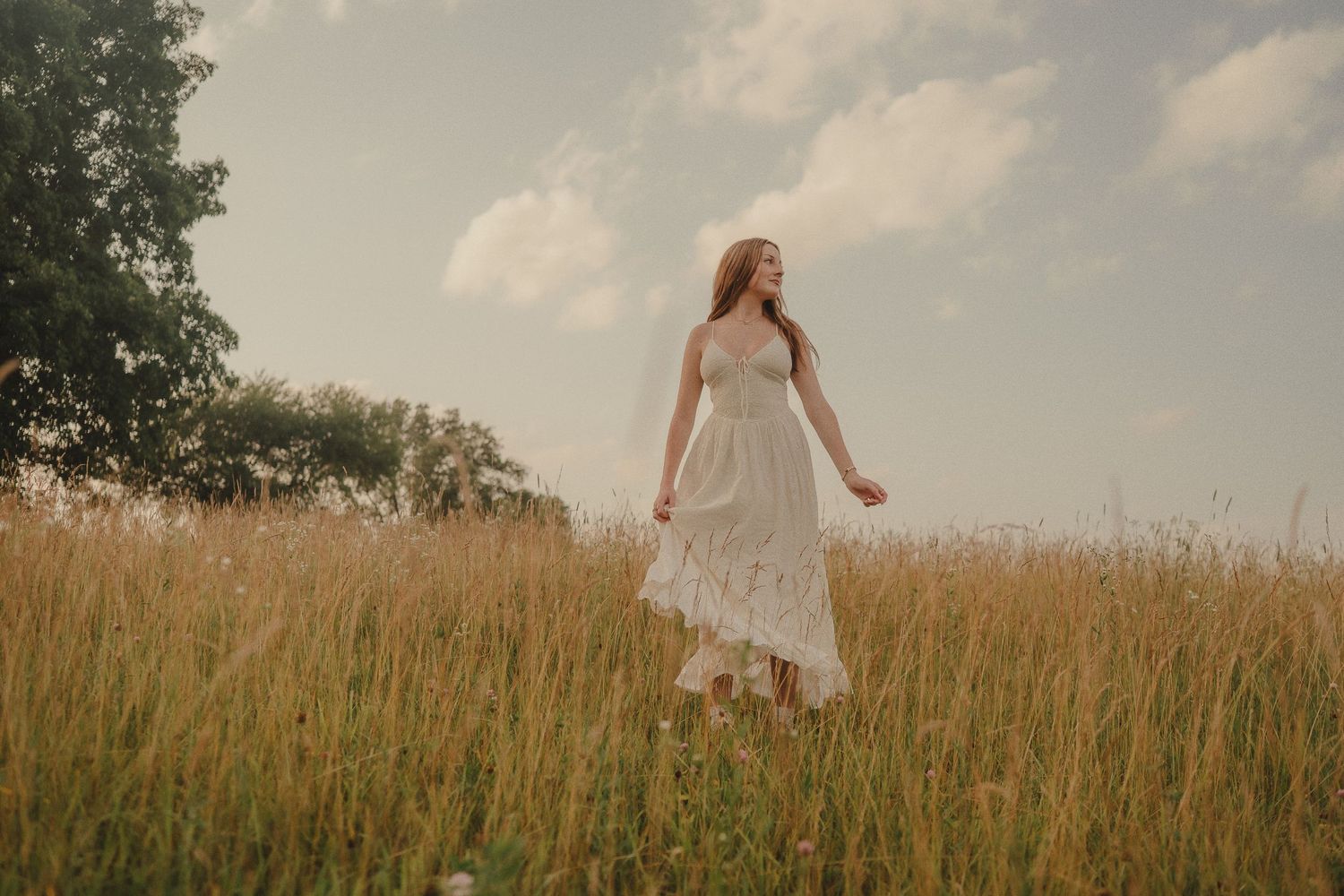 A young woman in a white lace dress walks gracefully through a golden meadow under a partly cloudy sky.