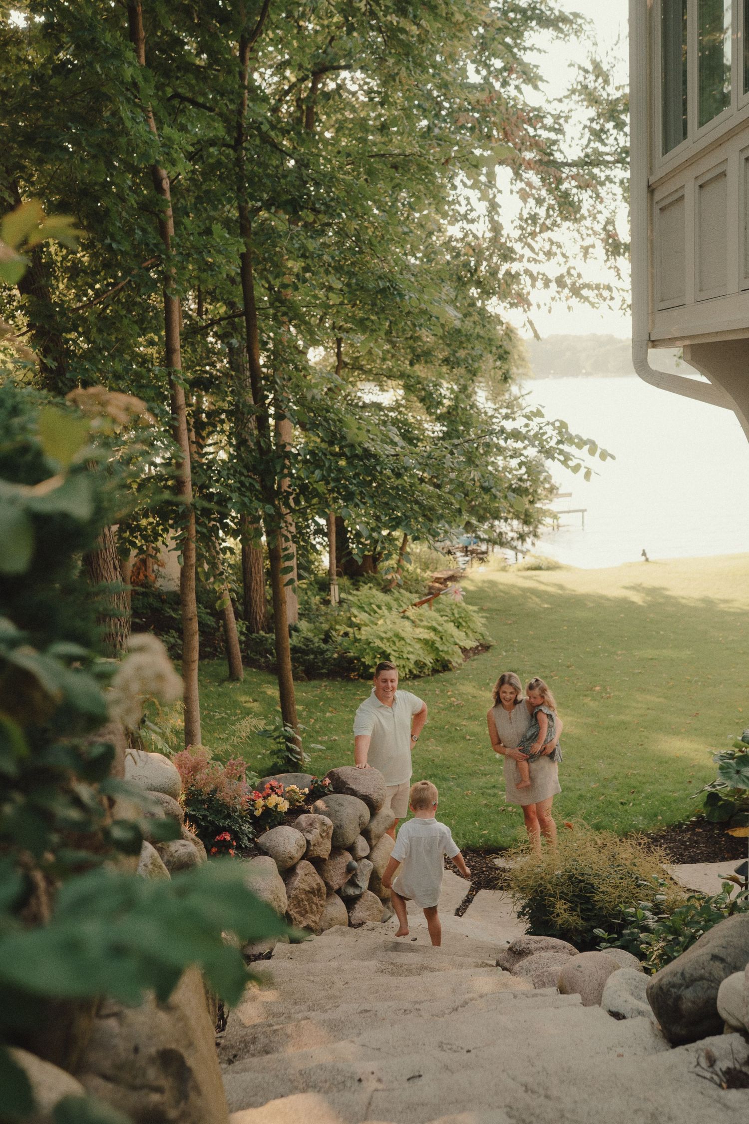 Family walking up stone steps toward a lakeside home surrounded by lush green trees and gardens at golden hour.