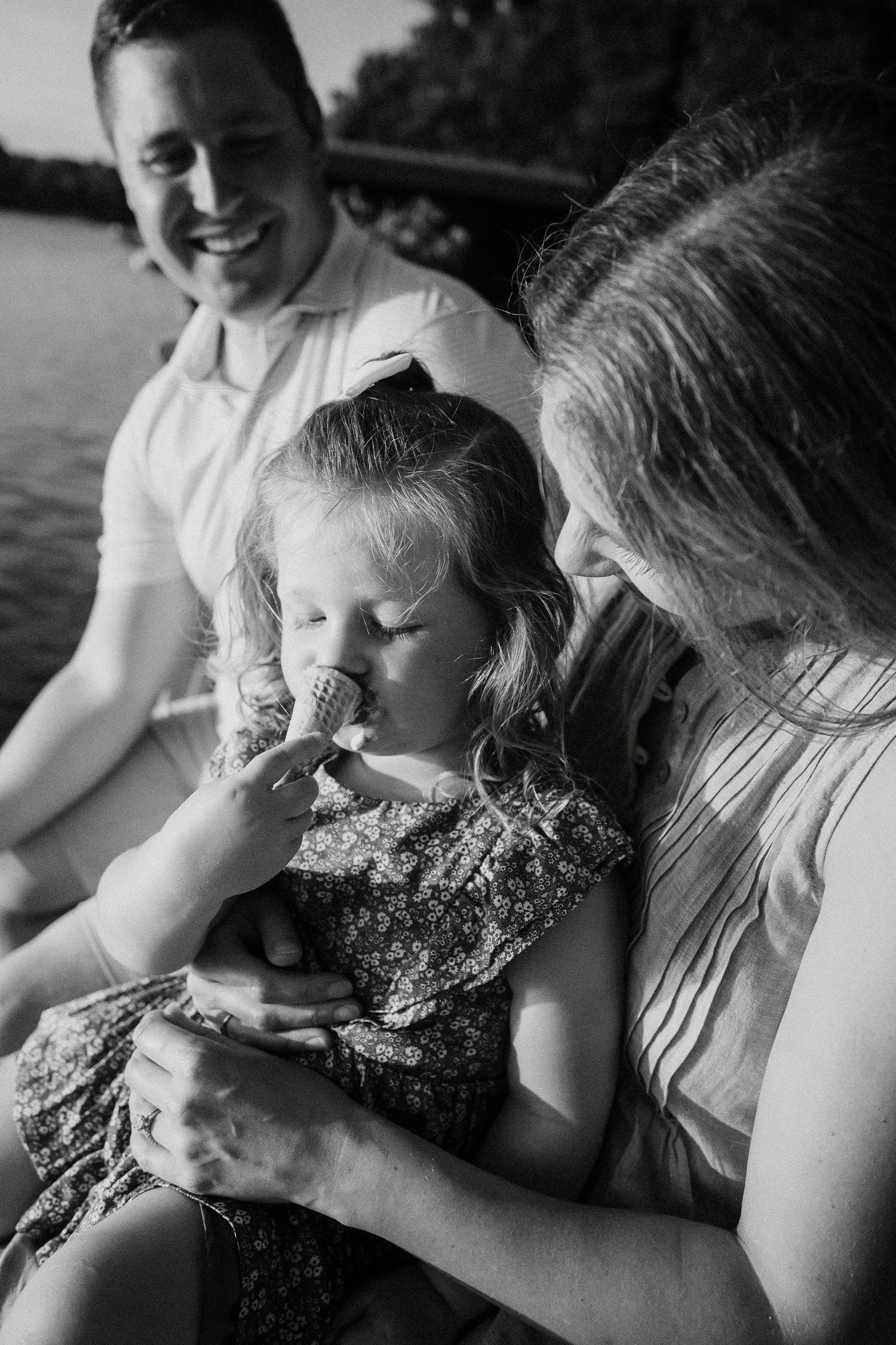 Black and white photo of a smiling family with a young girl eating an ice cream cone near a lake.