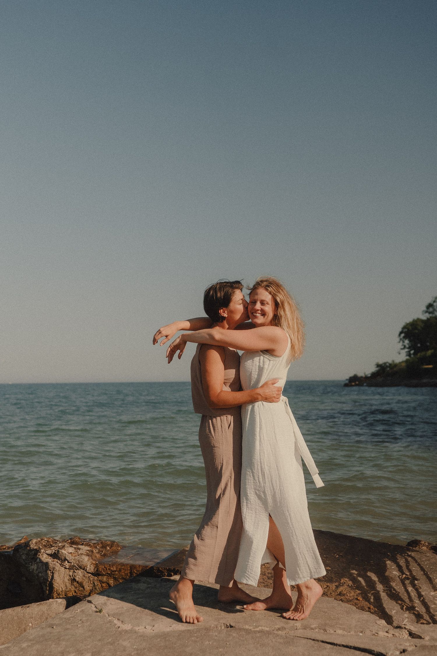 Two women in light dresses embrace and laugh together on rocky shoreline with calm blue ocean backdrop.
