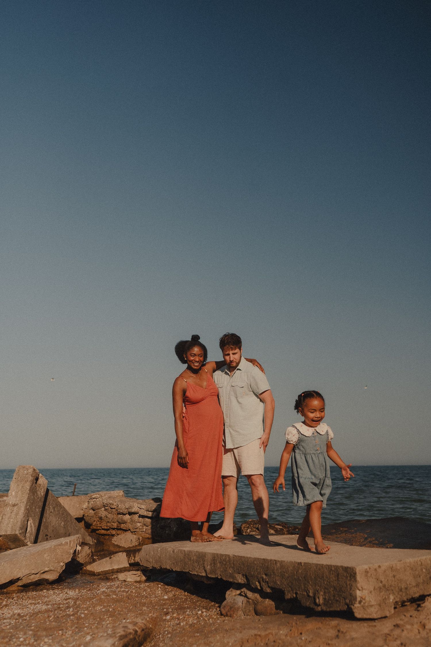 A multiracial family of three poses on rocky shoreline with calm ocean and clear blue sky behind them.