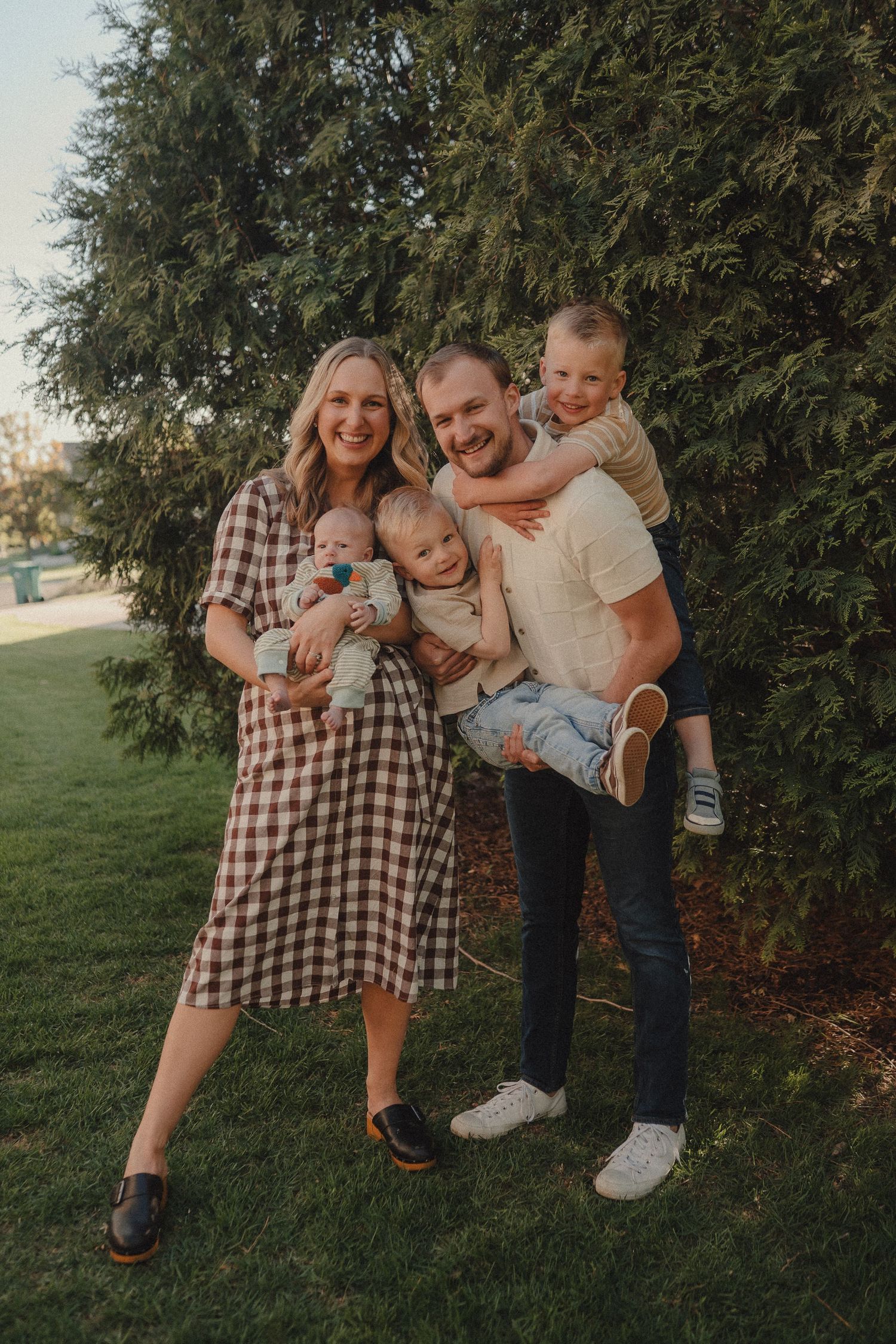 Happy family of four posing outdoors near evergreen trees, parents holding two young children on a sunny day.