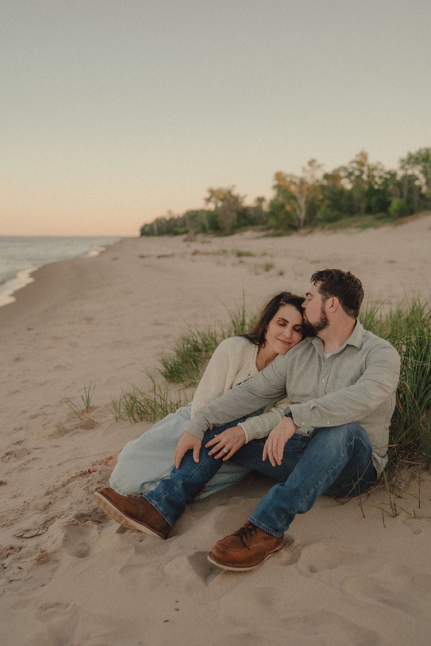 Man kisses woman on cheek as they sit together on sandy beach at sunset.