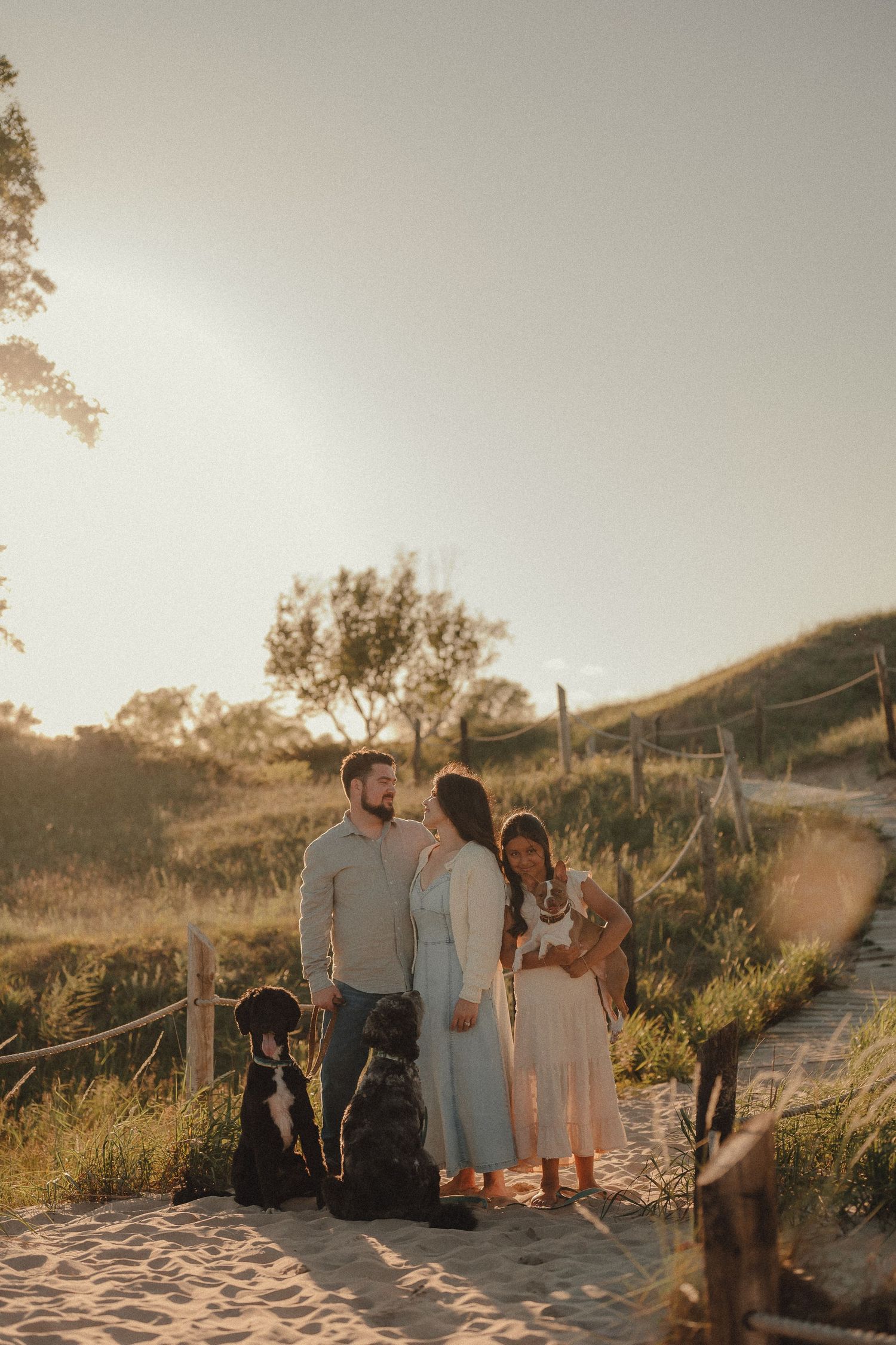 Couple with two black dogs standing on sandy path at golden hour with lush green hills in background.