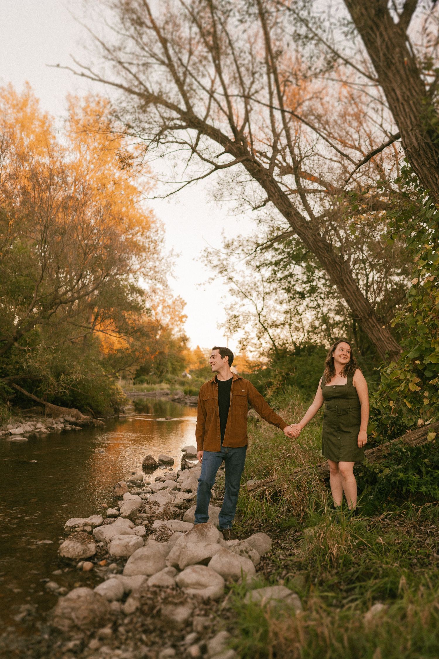 A couple stands together on a rocky riverbank path during sunset with fall foliage in background.
