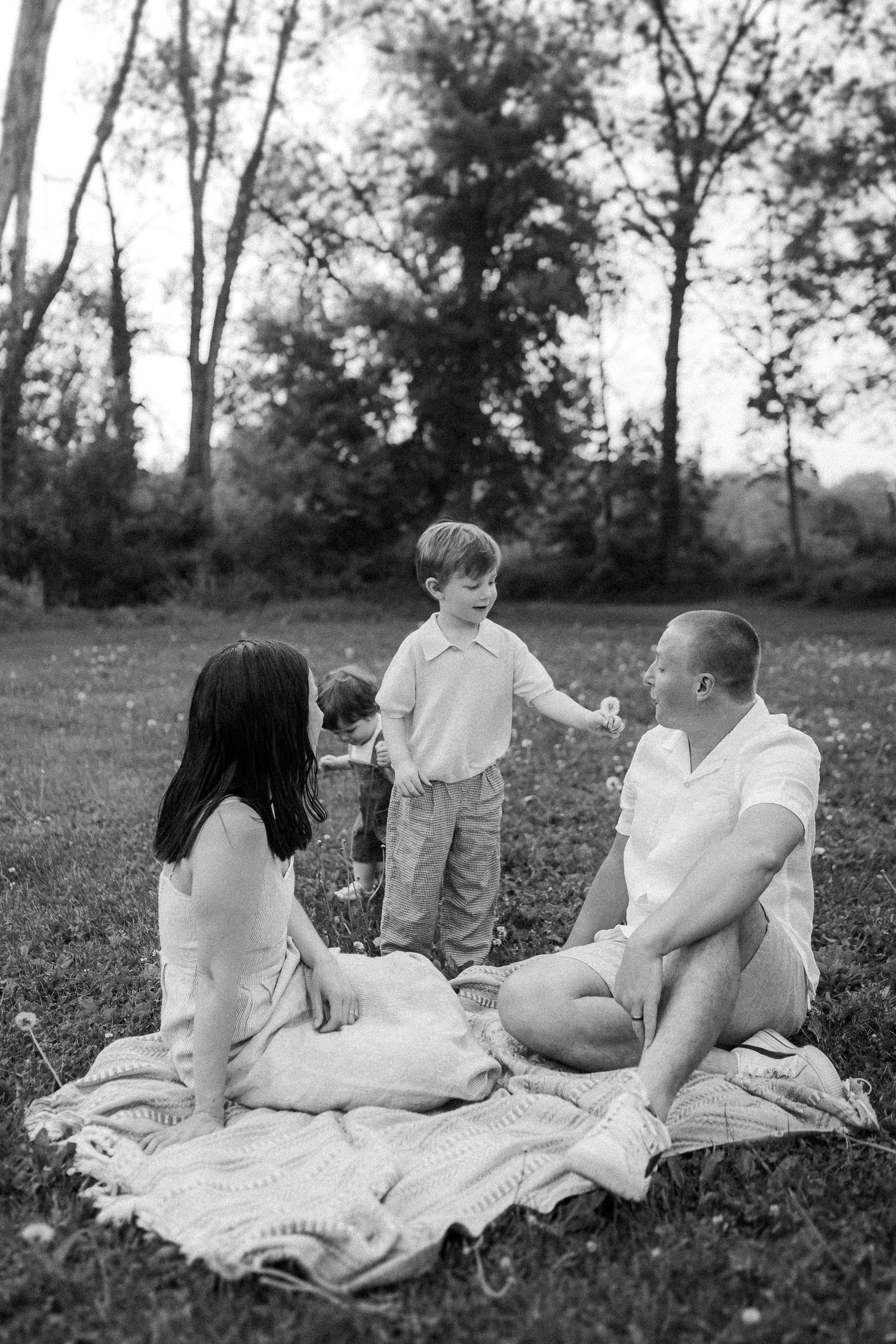 A candid black and white photo of a family having a picnic on a blanket in a grassy field surrounded by trees.