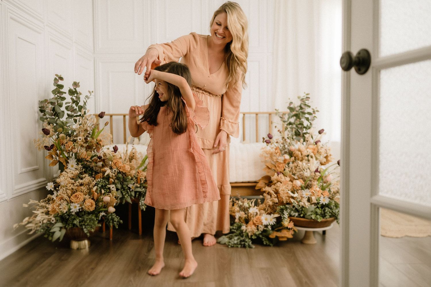 A figure in a flowing peach dress stands in front of floral arrangements on wooden floors in a bright interior setting.