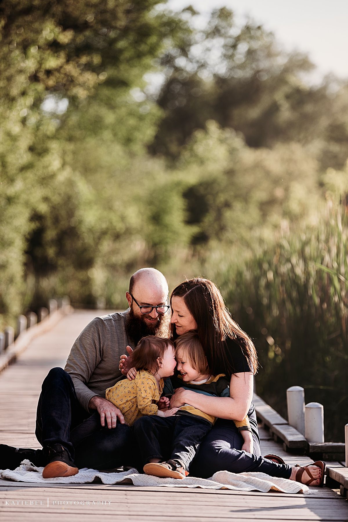 Family shares intimate moment together while sitting on wooden boardwalk surrounded by nature.