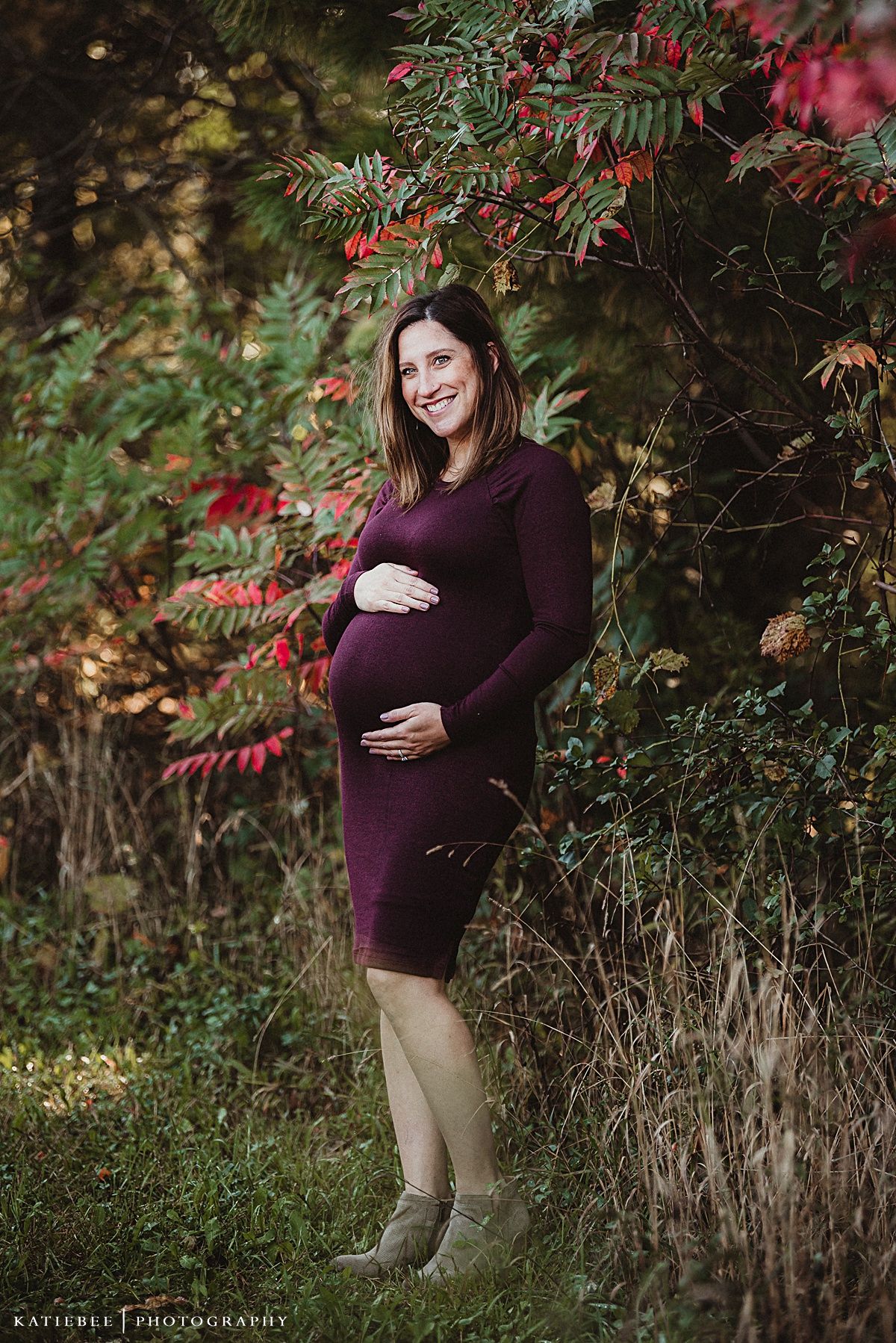 An expectant mother in a burgundy dress poses under red autumn foliage.