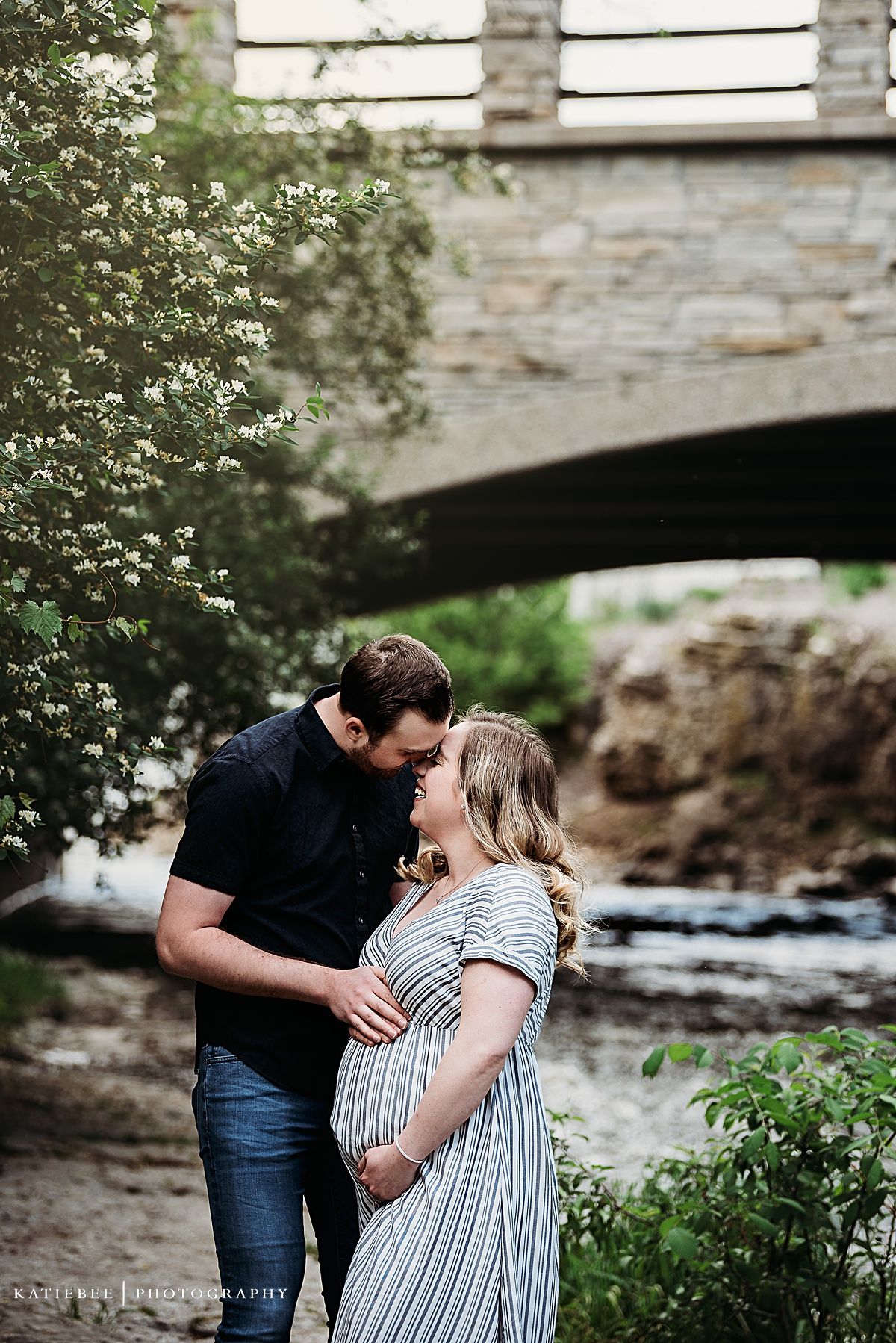 A romantic maternity photo under a stone bridge with flowing water in the background.