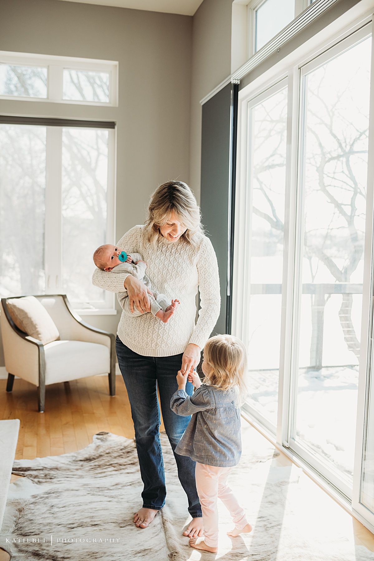 Mother and toddler stand by bright windows holding newborn in cable knit sweater.