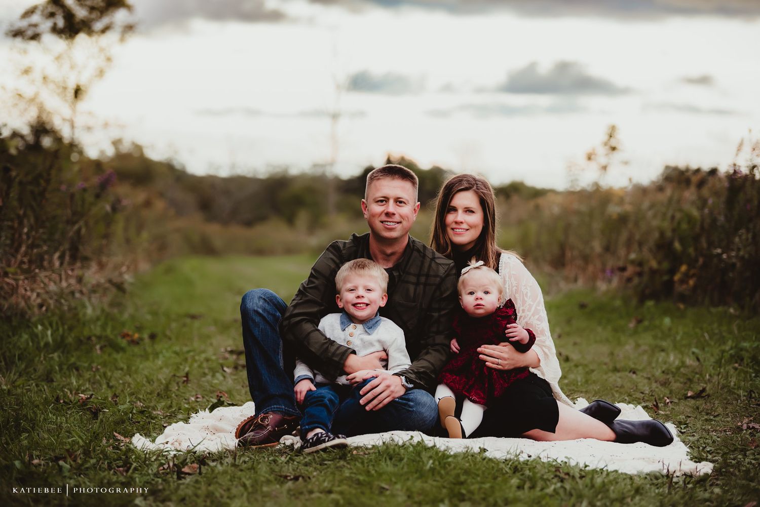 A relaxed family portrait on a white blanket surrounded by natural countryside scenery.