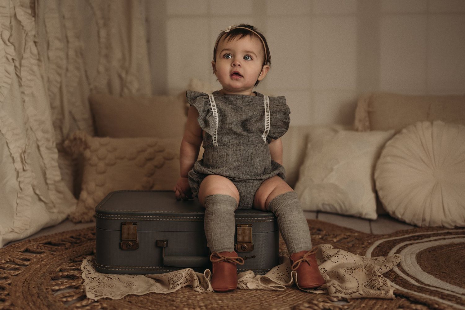 Baby in grey romper and knee socks sits on vintage suitcase.