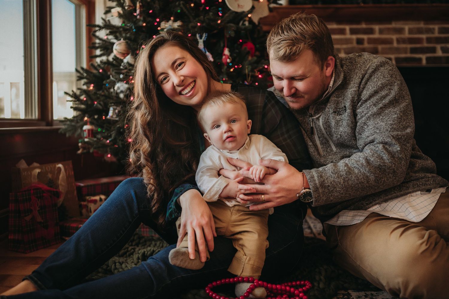 Candid family portrait by Christmas tree with exposed brick and natural window lighting.