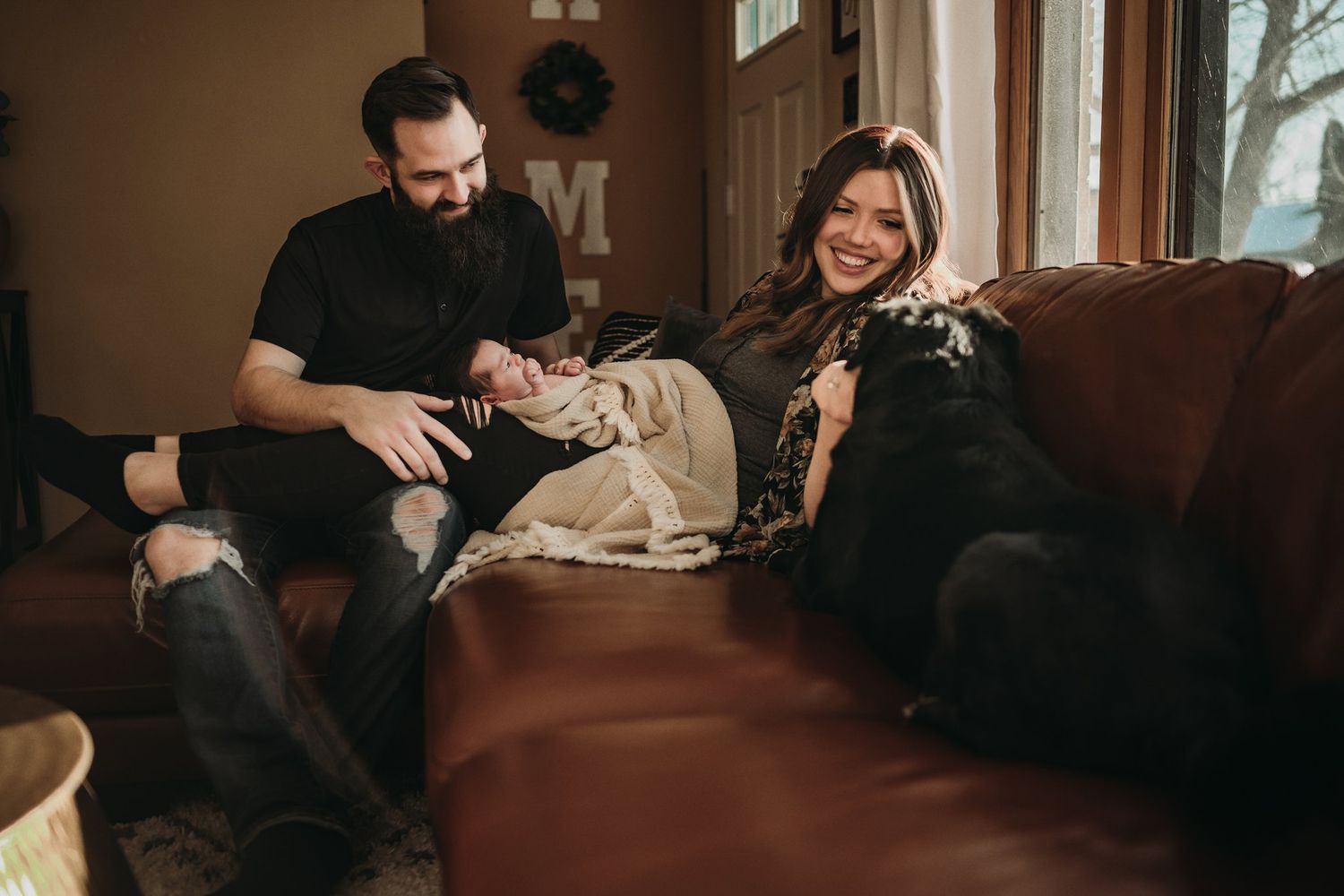 Family relaxing on brown leather couch with infant during intimate home photoshoot.