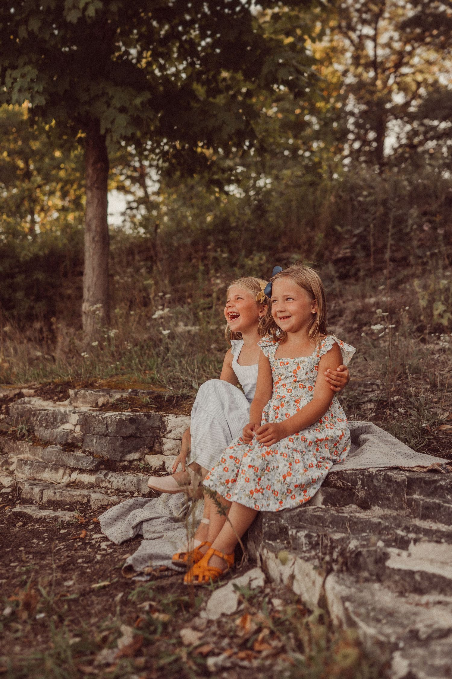 Two children laugh while sitting together on a rock formation in a wooded setting.