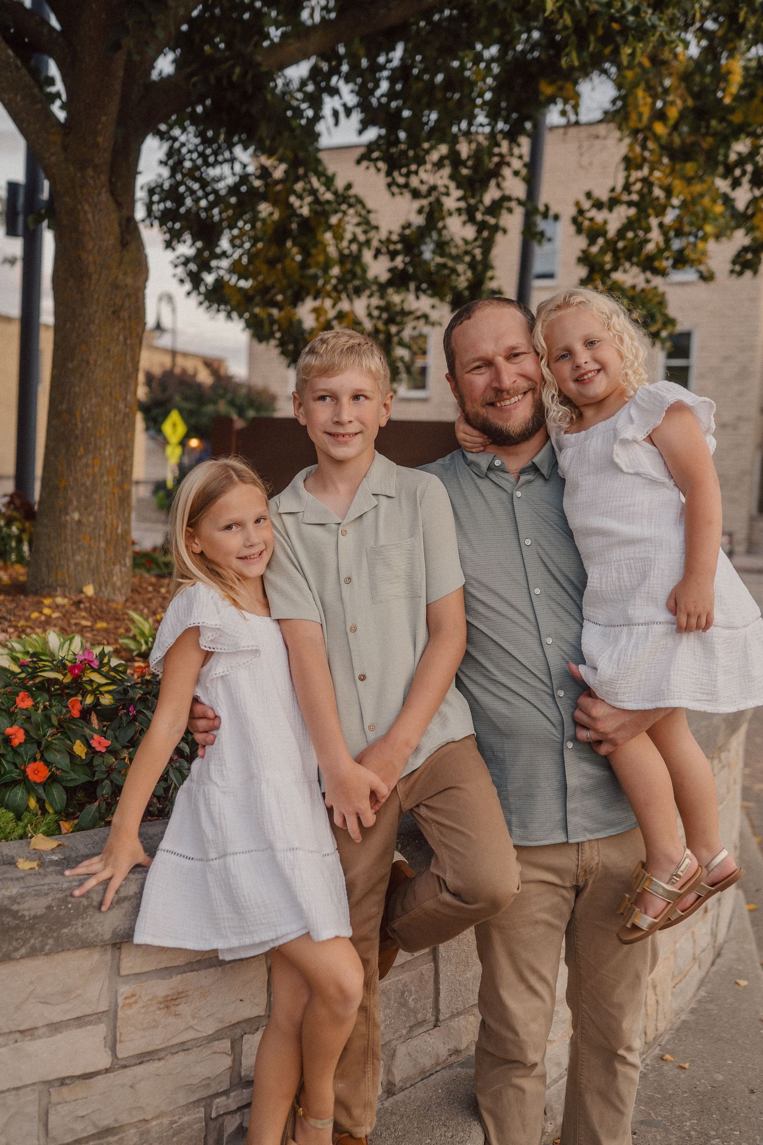 A family portrait taken outdoors against a stone wall with a large tree and flowers in the background.