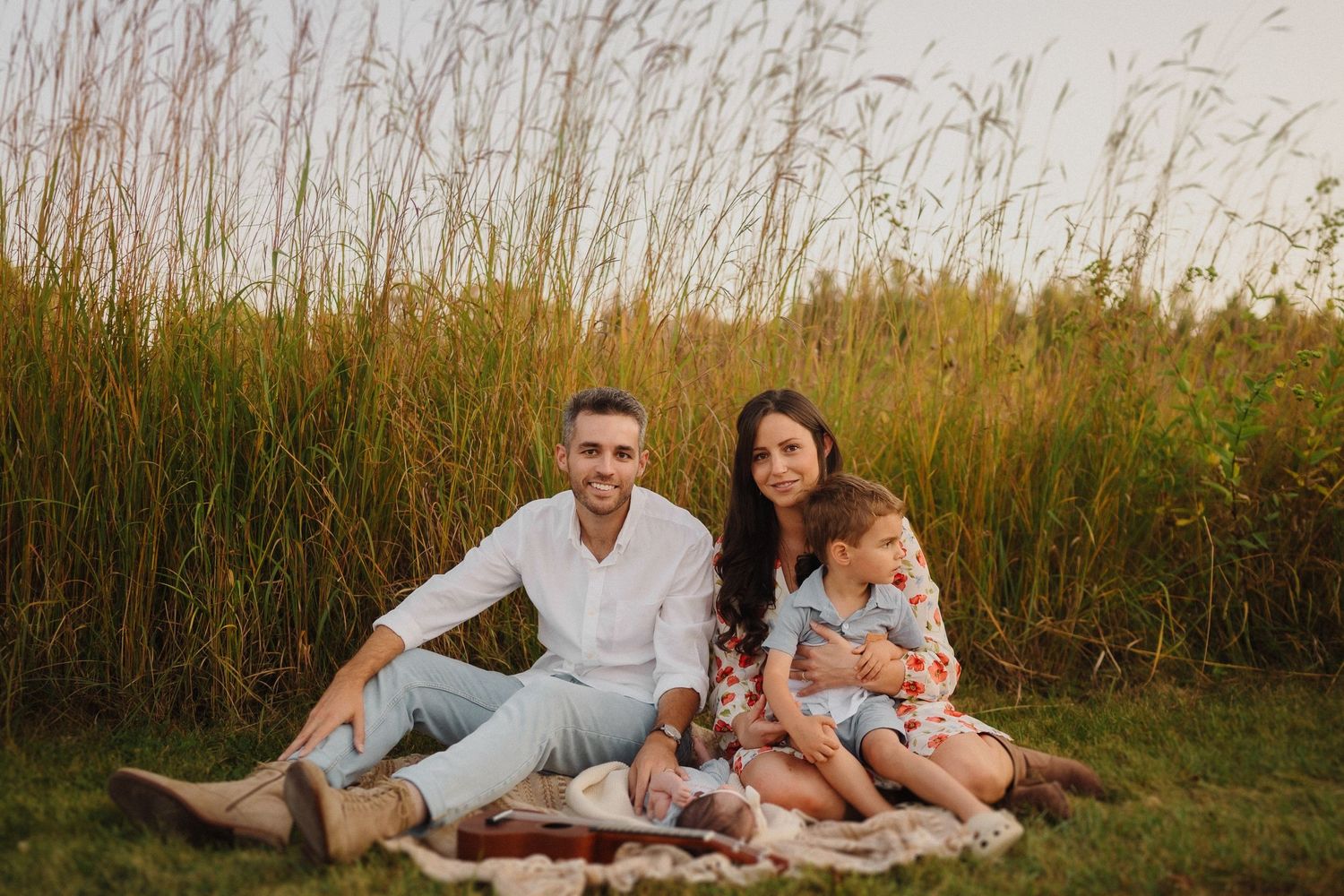 Family relaxing on picnic blanket against backdrop of tall grass at sunset.