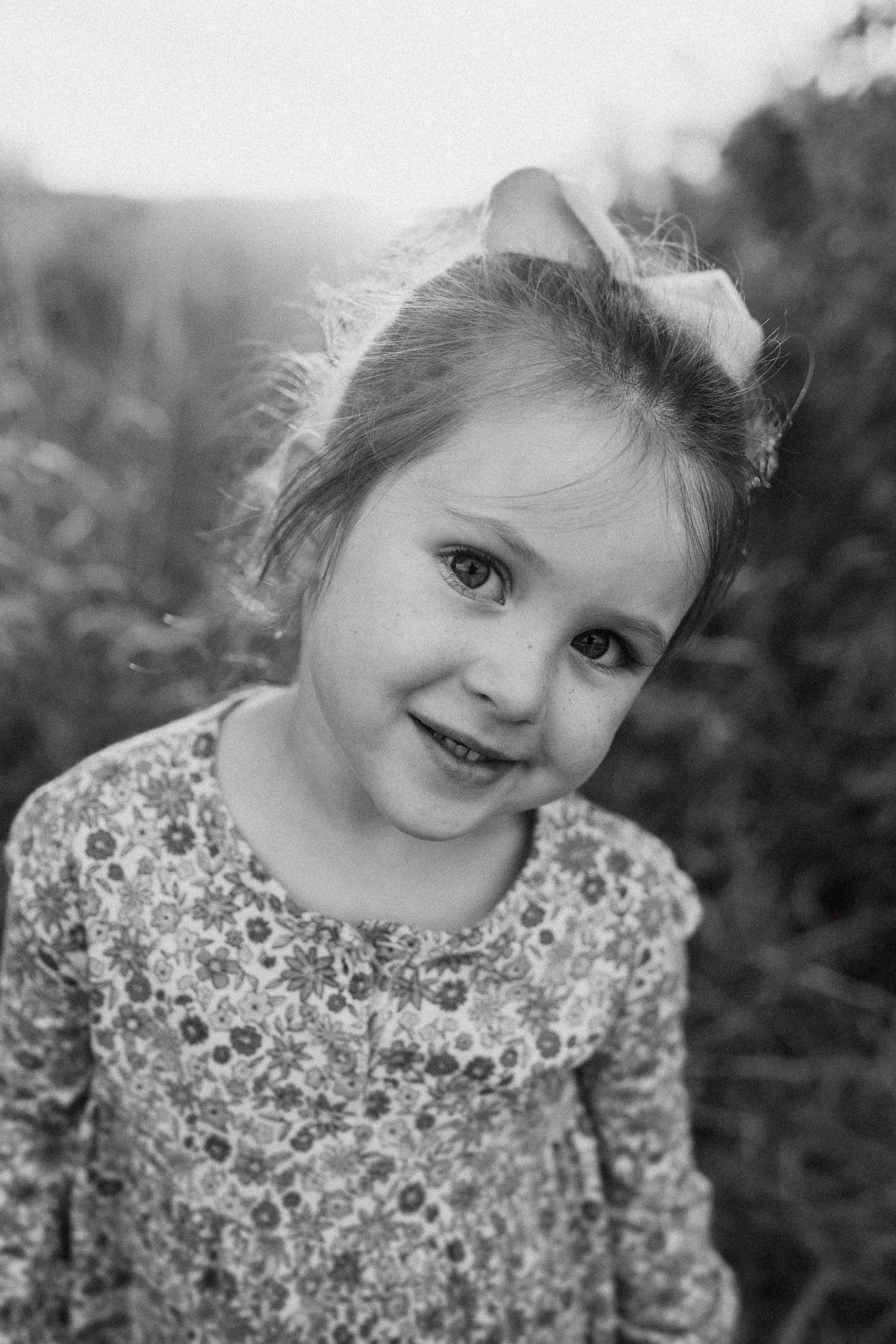 Black and white portrait of young child wearing floral print dress and hair bow smiling at camera.