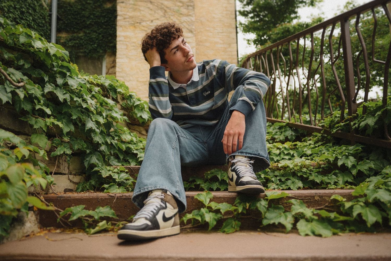 Someone in a striped sweatshirt and jeans sits pensively on ivy-covered stone steps while wearing trendy sneakers.