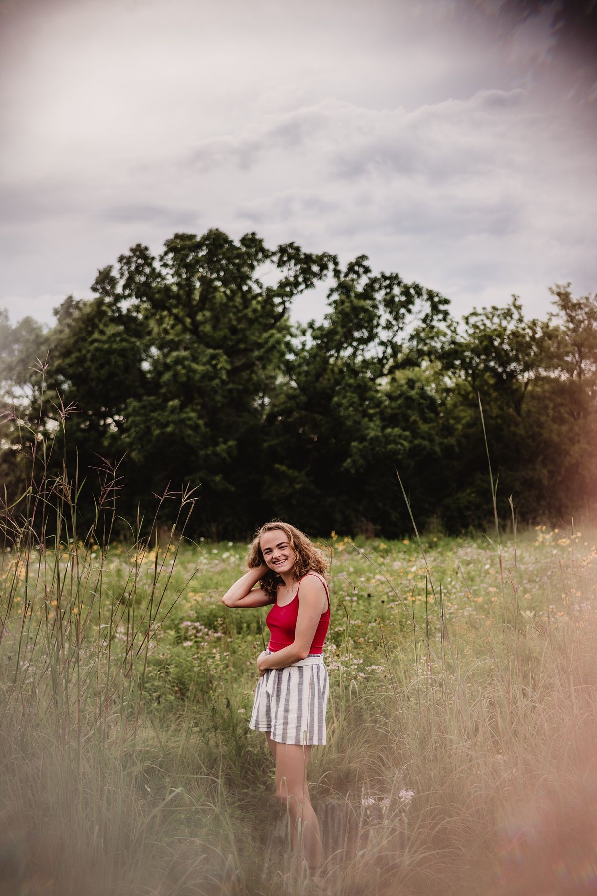 Summer portrait in a meadow with tall grass and trees, featuring a red top and striped shorts outfit.