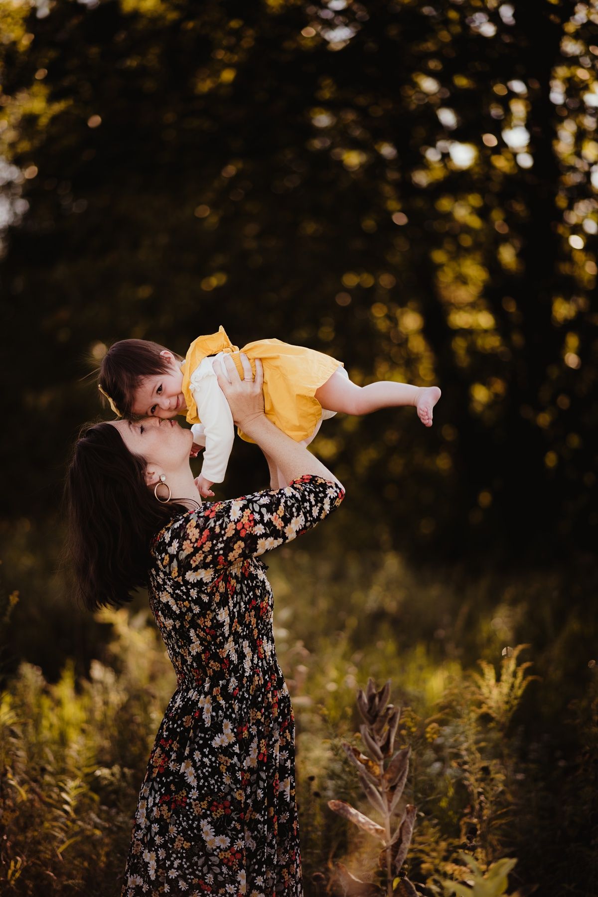 Playful outdoor portrait with adult lifting baby in yellow dress against sunset backdrop.