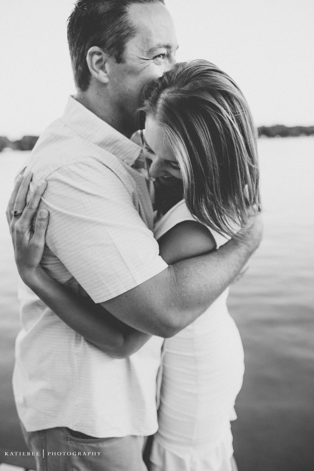 An intimate black and white portrait of a couple embracing by the water.