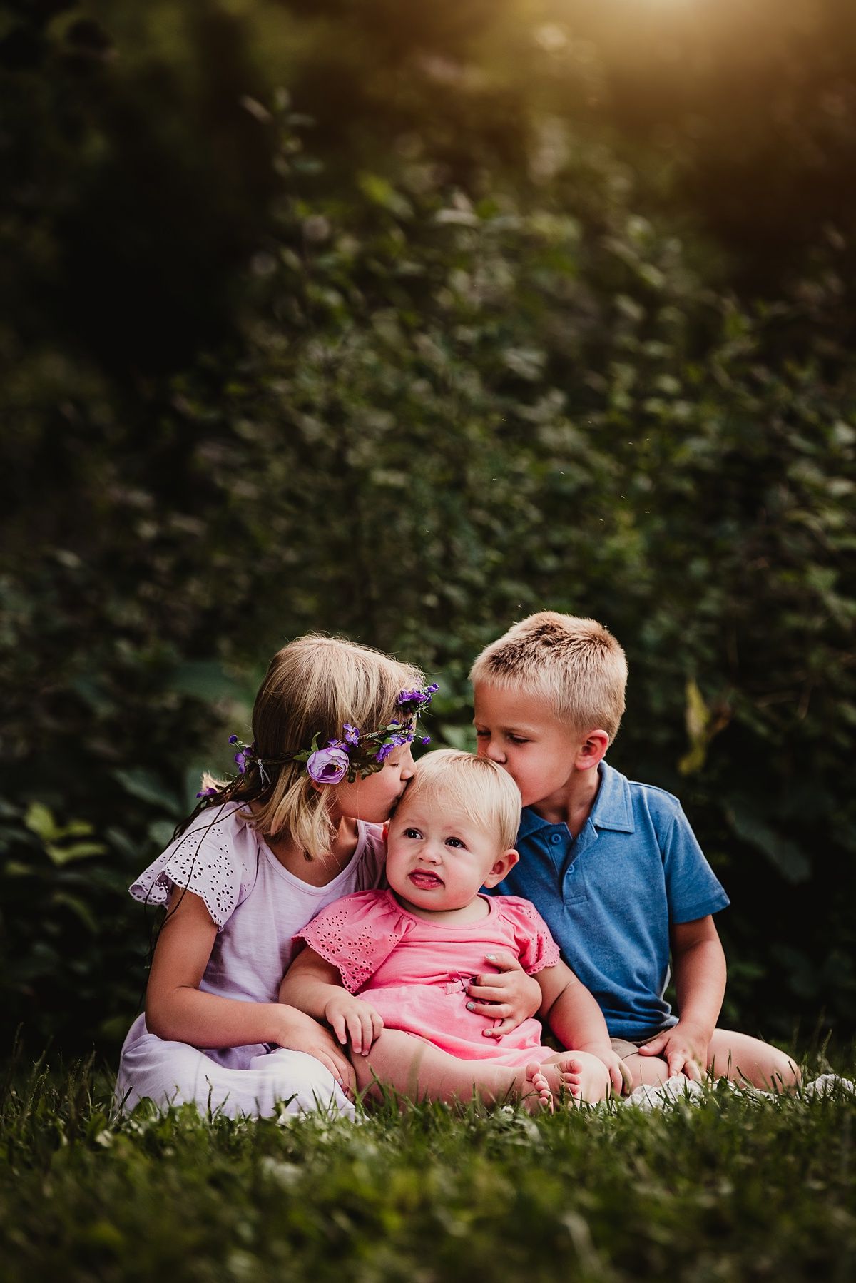 Three children sitting together on grass in a garden with soft evening lighting.