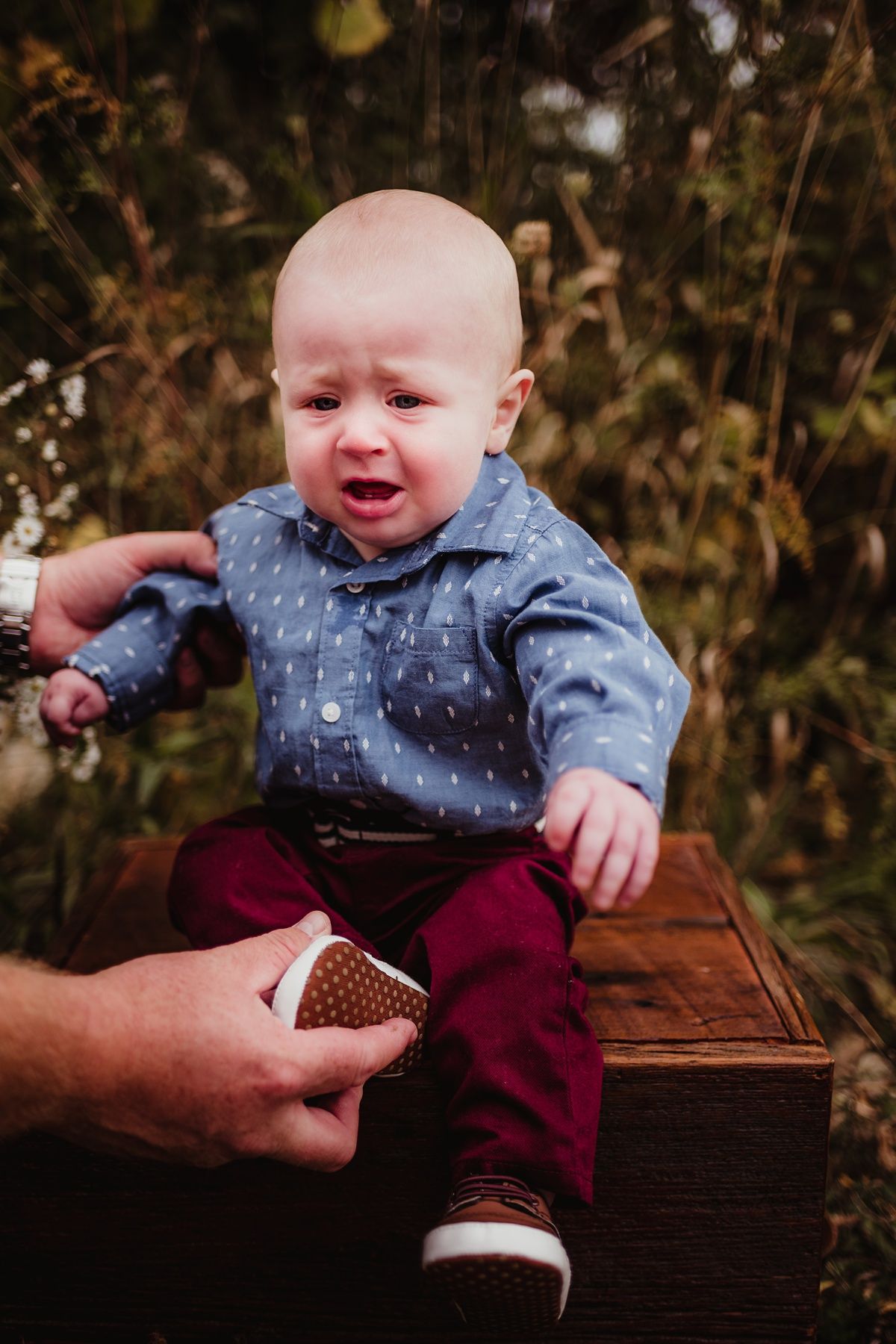 Small child in polka dot shirt and burgundy pants sits crying on wooden surface.