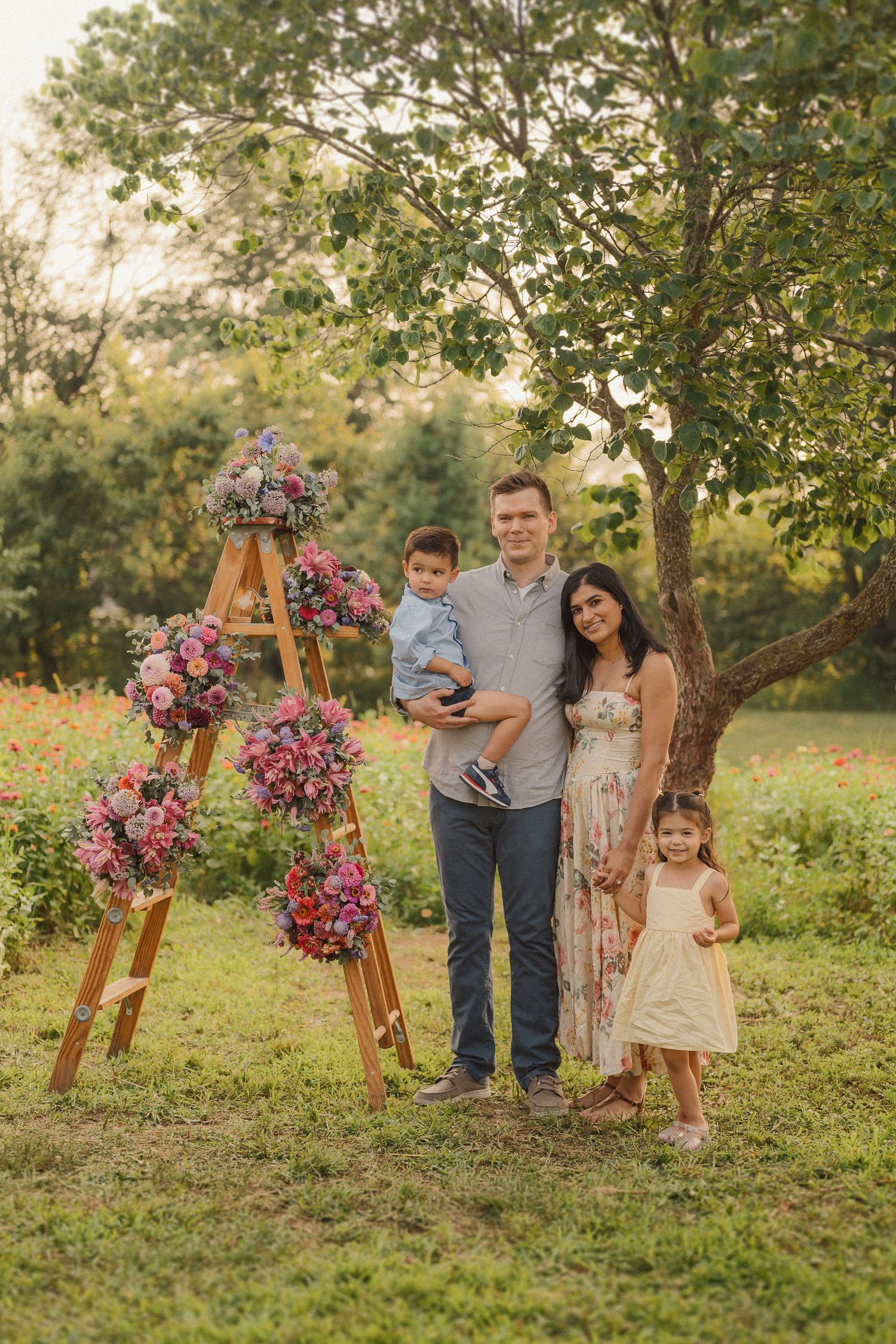 A family poses together outdoors near a wooden ladder decorated with pink and purple floral arrangements.