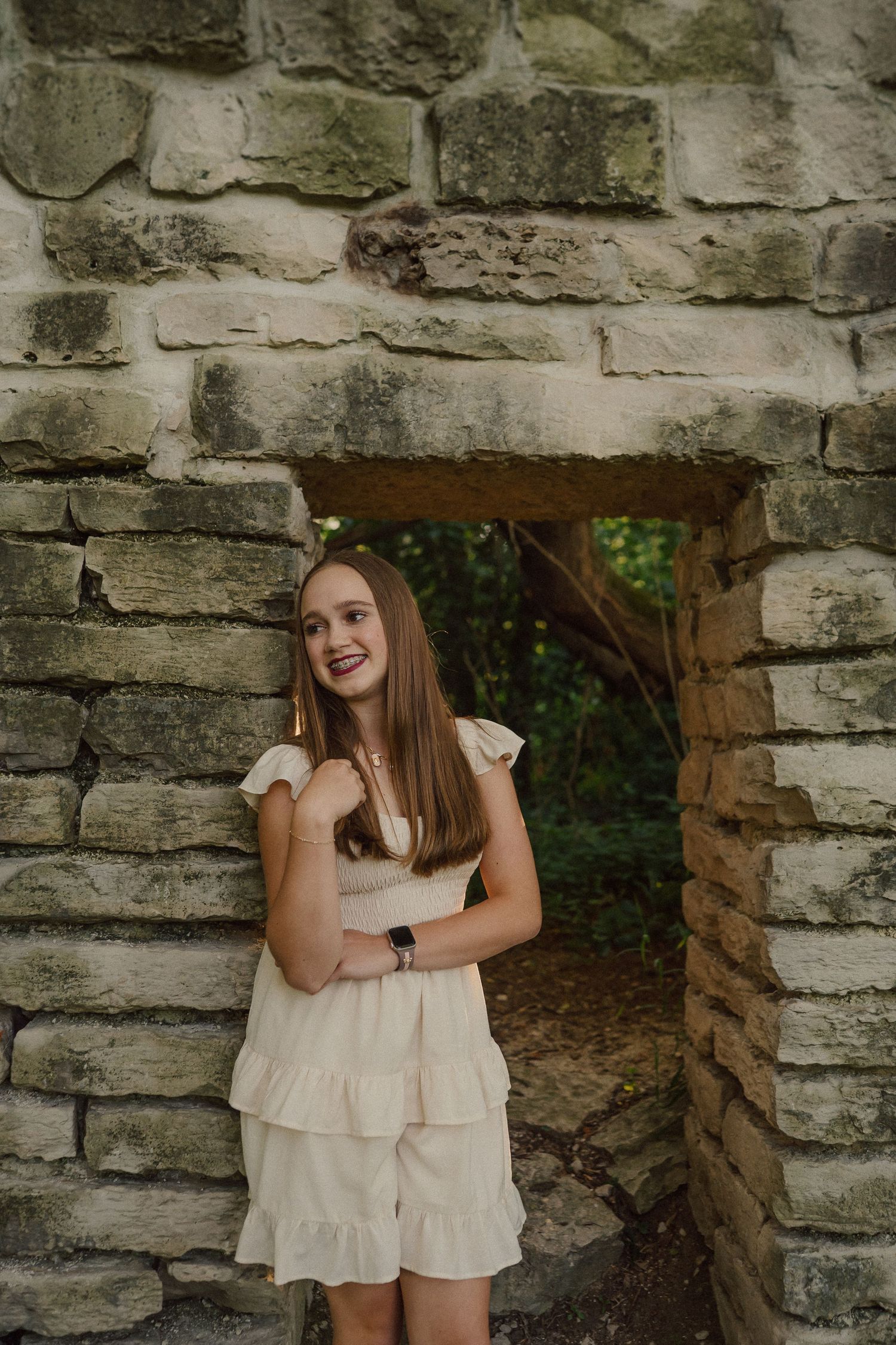 A person in a cream-colored ruffled outfit poses playfully against a rustic stone wall.