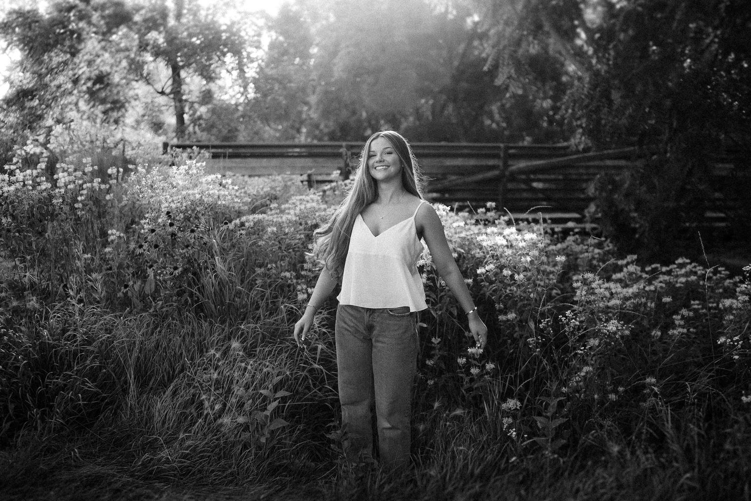 Black and white portrait of a person standing in a wildflower garden with sunlight streaming through trees.