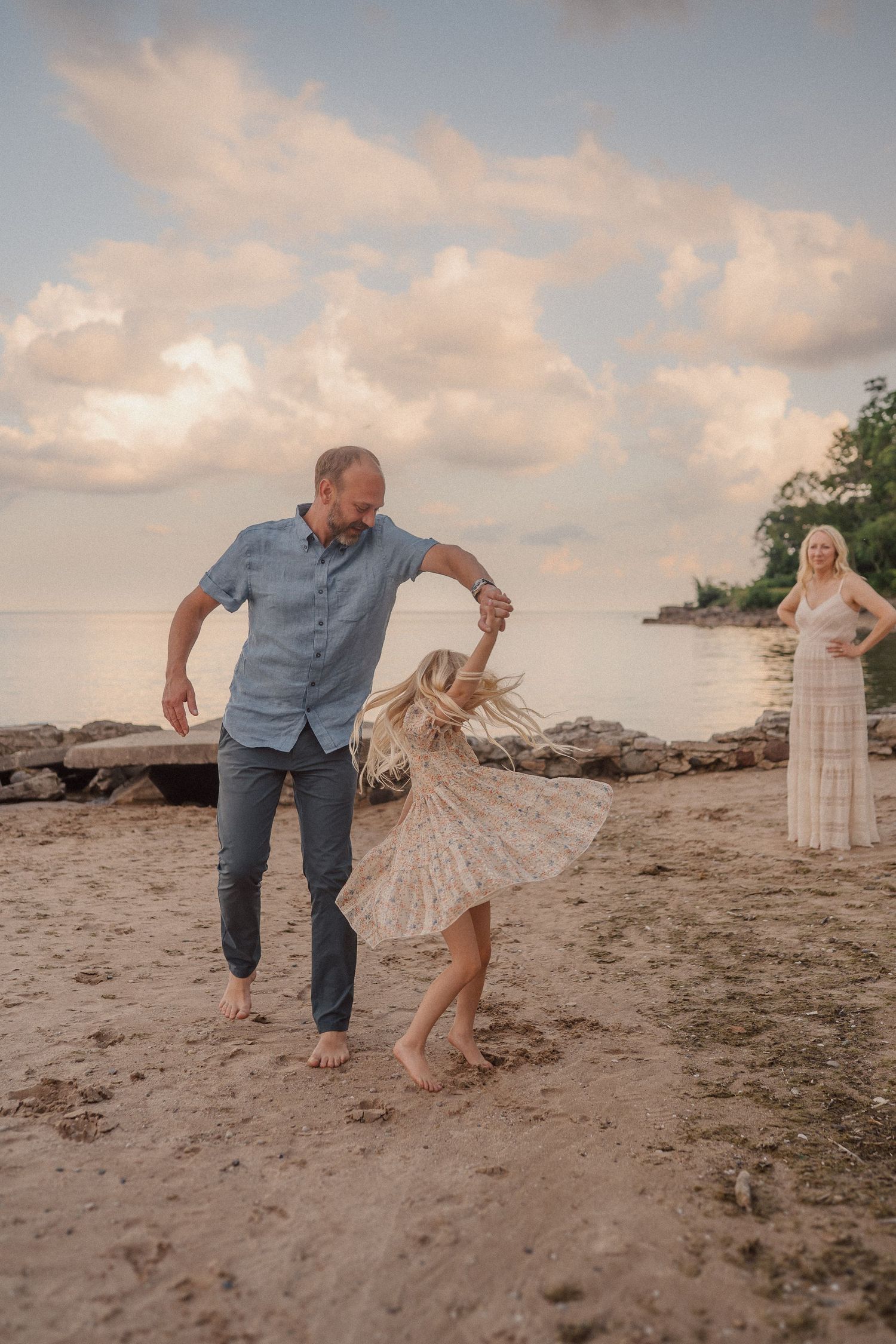 A family dancing together on a beach at sunset with scenic clouds and driftwood in the background.
