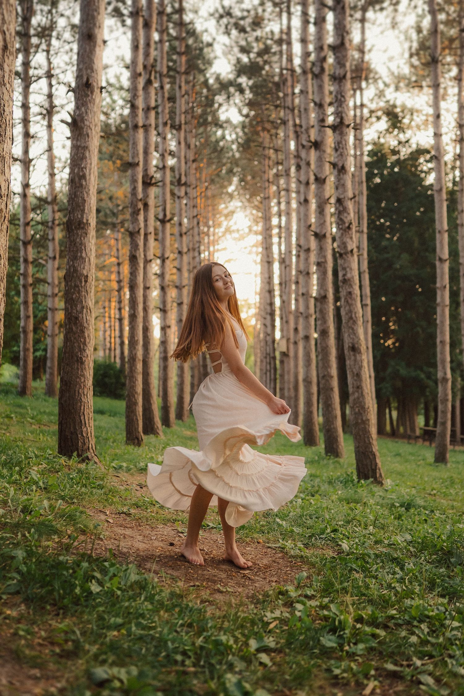 A flowing white dress twirls through a sun-dappled pine forest path during golden hour.