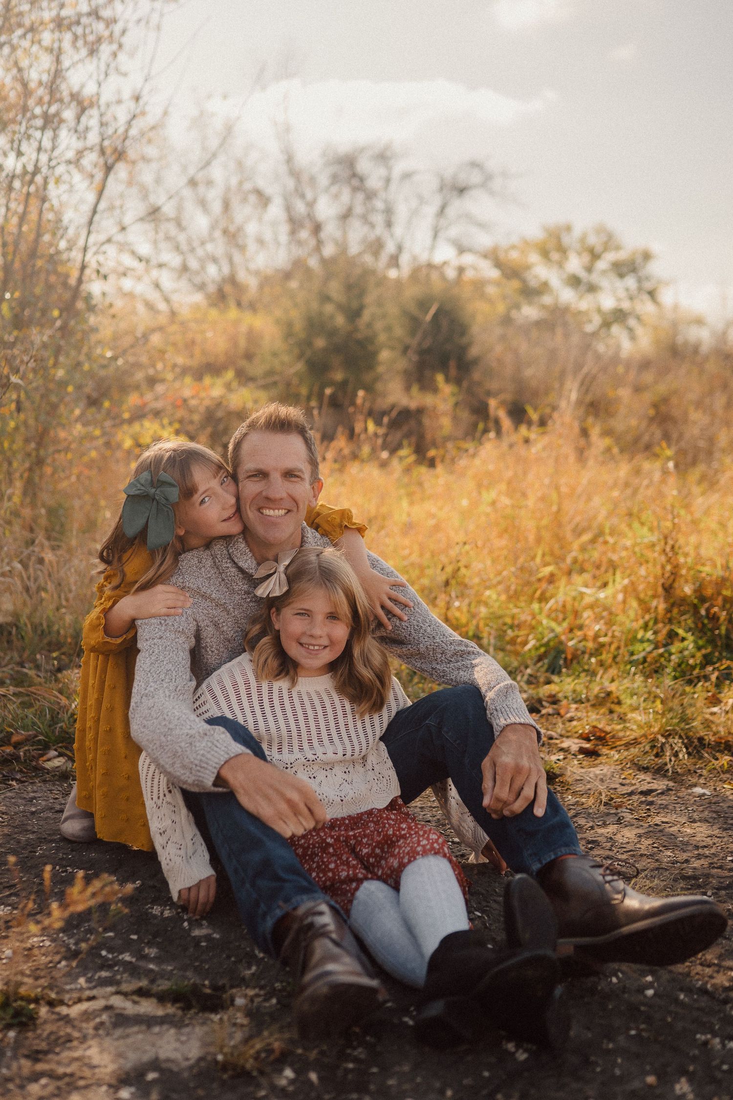 A couple sits together on the ground during a golden autumn photoshoot in a natural outdoor setting.
